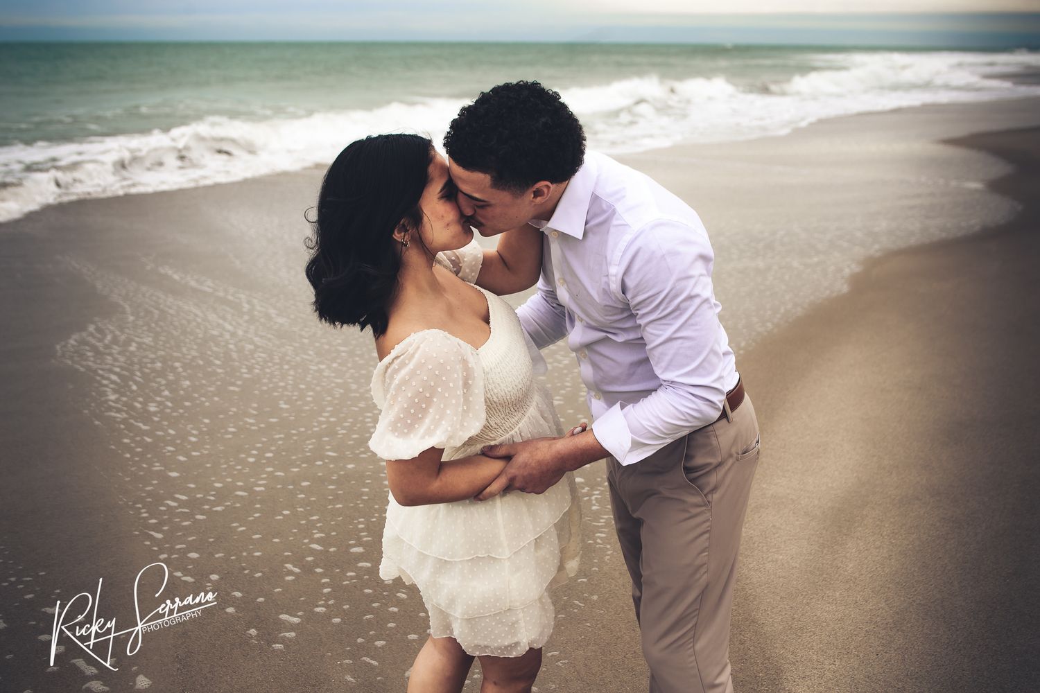 A romantic couple shares a tender moment on a sandy beach with ocean waves in the background.