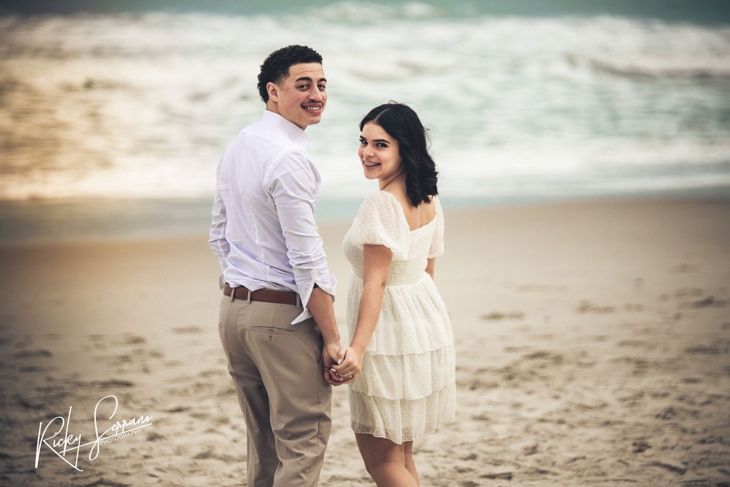 A couple stands holding hands on a peaceful beach at sunset, dressed in light romantic attire.