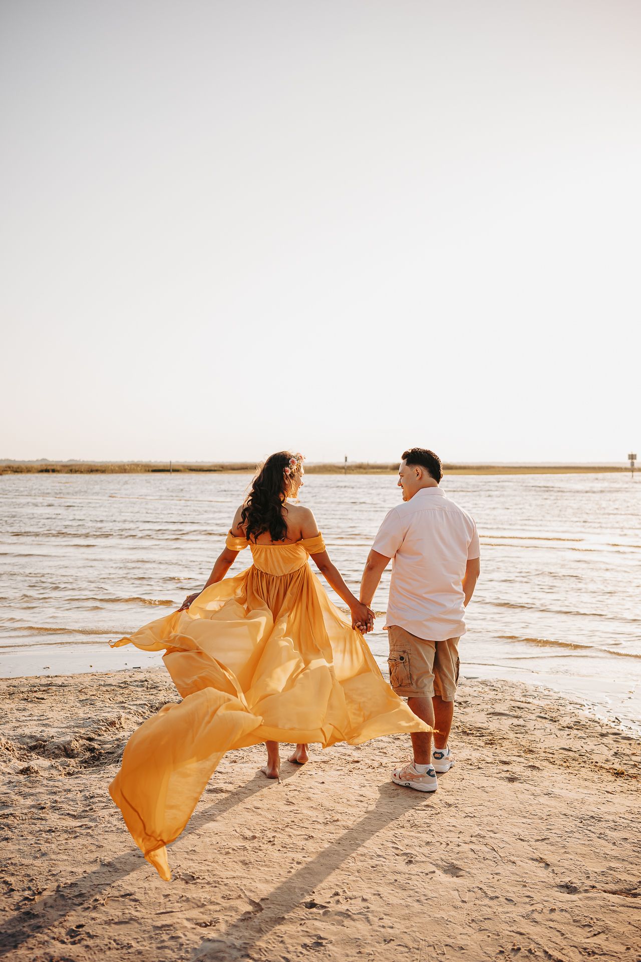 A couple walking together on a beach at sunset as a flowing yellow dress catches the wind along the shore.