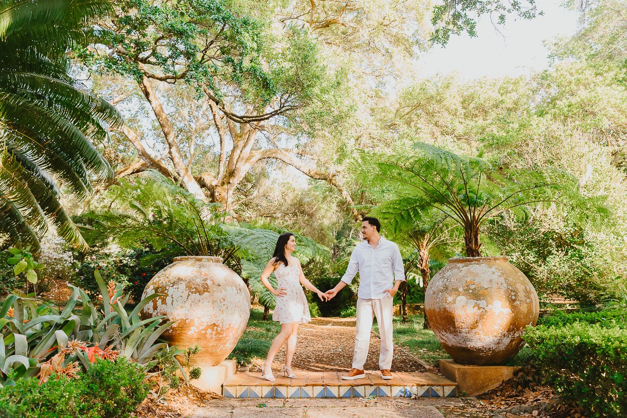 Couple in white outfits standing between large stone spheres on garden steps surrounded by lush tropical foliage.