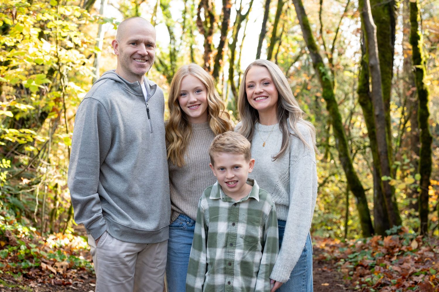 Family portrait of four people smiling together in casual attire among autumn trees.