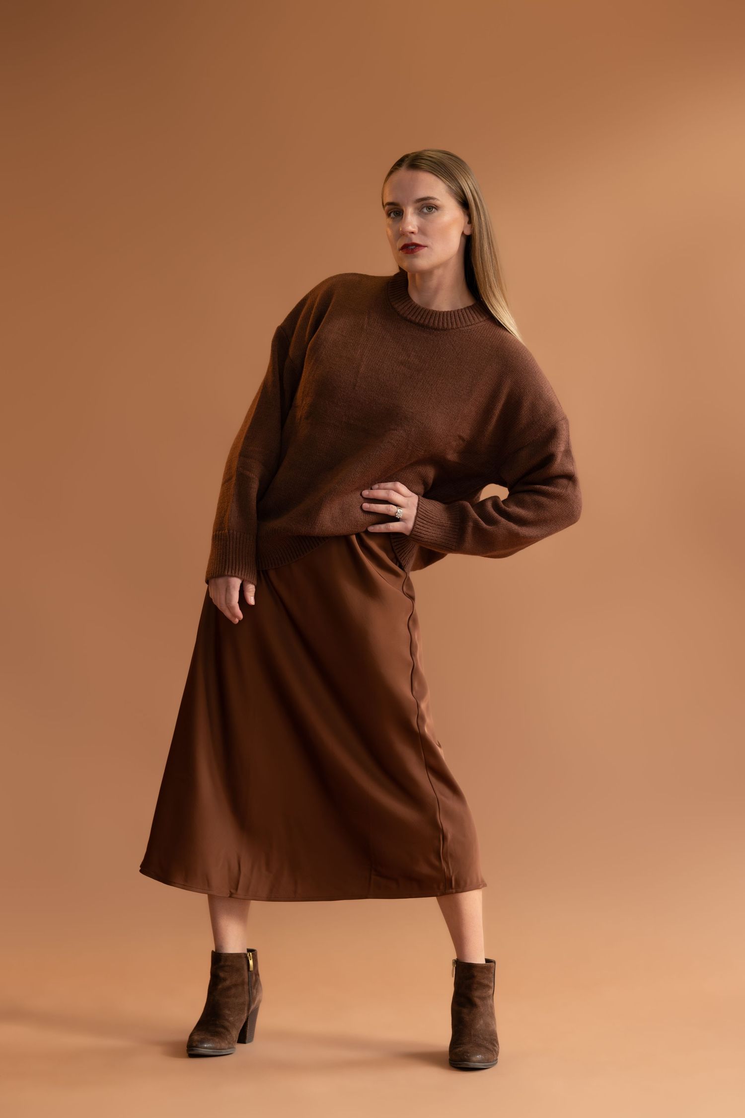 Model in brown sweater dress and boots posing against warm-toned studio background.