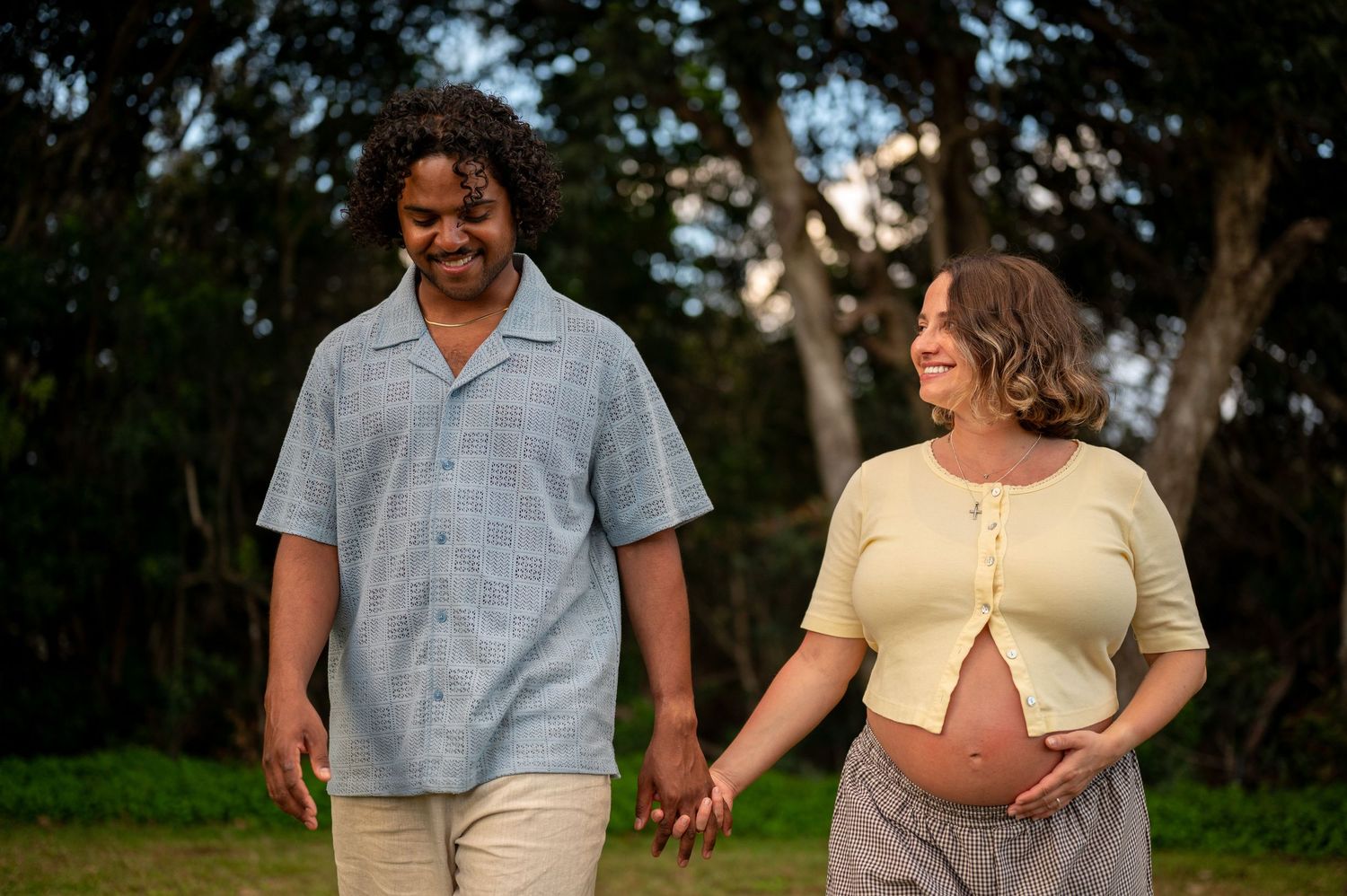 Pregnant couple holding hands and smiling while walking outdoors in a park with lush green trees in the background.