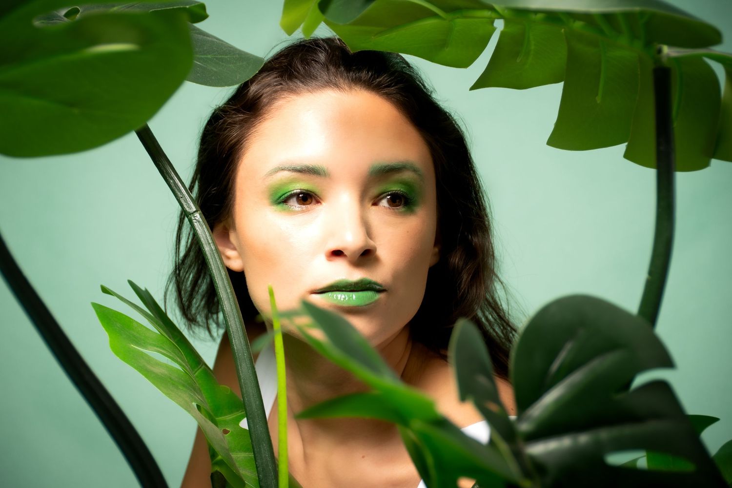 Creative portrait through monstera leaves with vibrant green makeup and dramatic lighting. Portland, Oregon. Studio photography.