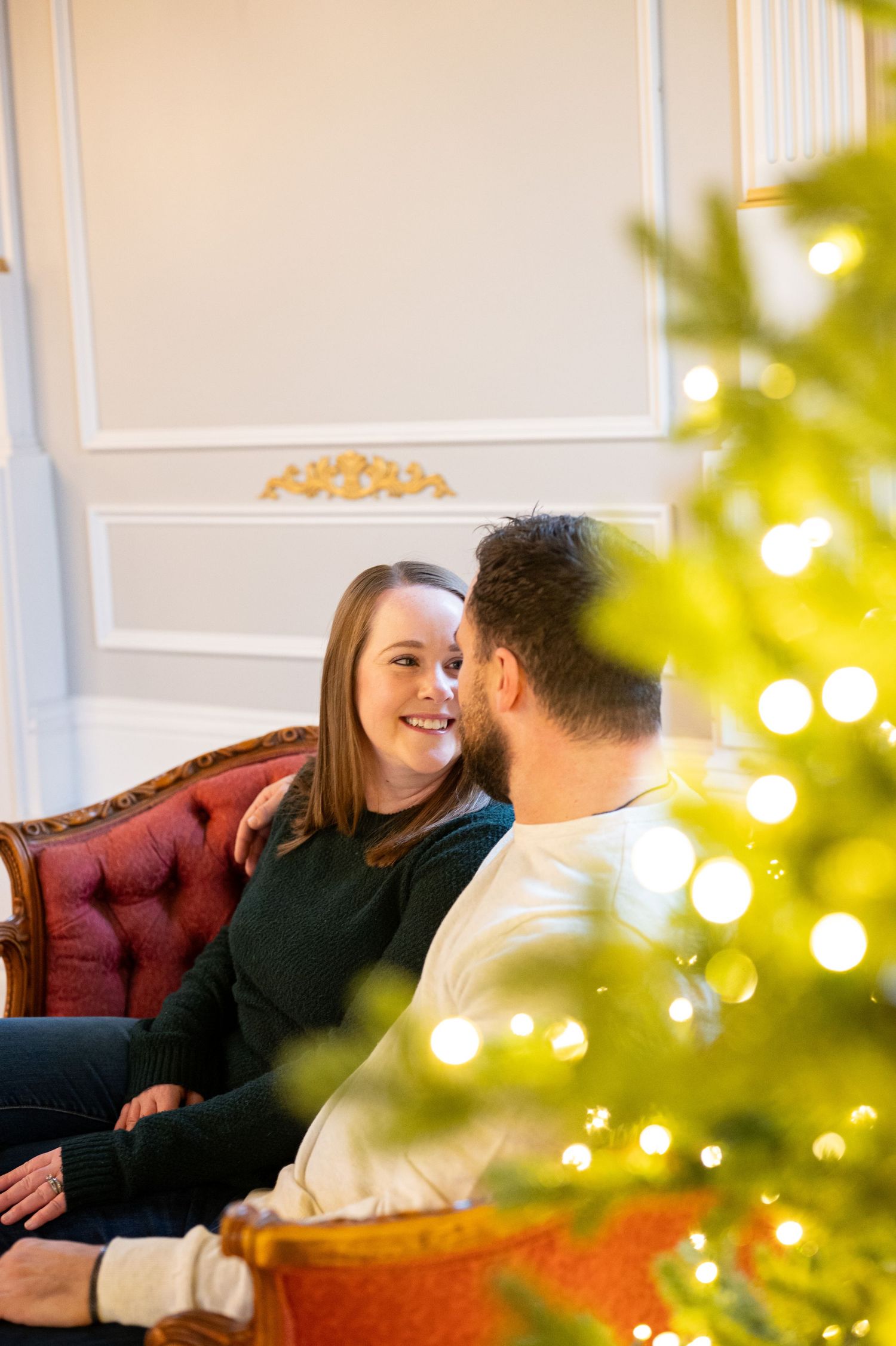 A couple shares an intimate moment on a red sofa with Christmas lights twinkling in the background. Family session. Portrait photography. Or