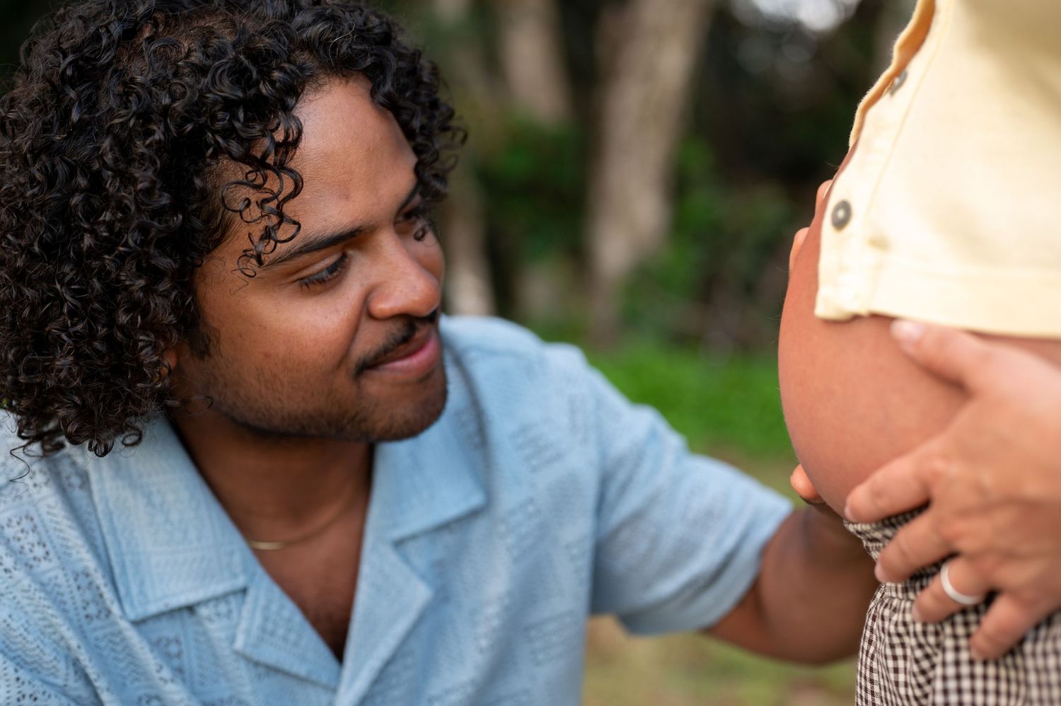 A man with curly hair tenderly touches a pregnant woman's bare belly outdoors among green trees.
