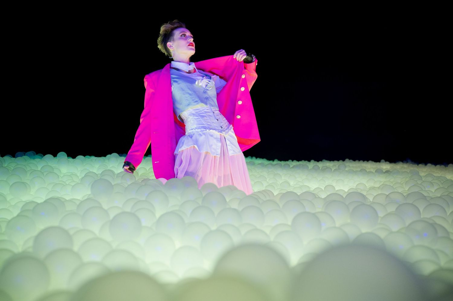 Figure in bright pink jacket poses against illuminated cloud-like backdrop. Hopscotch. Portrait Slam.