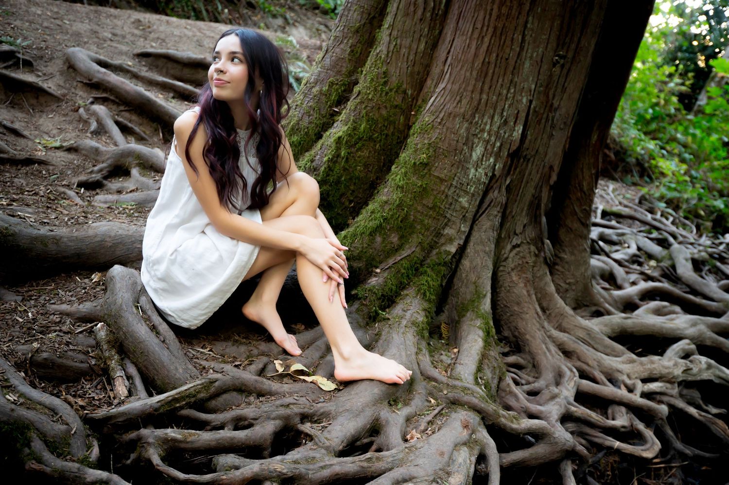 Person in white dress sitting among exposed tree roots in a forest.