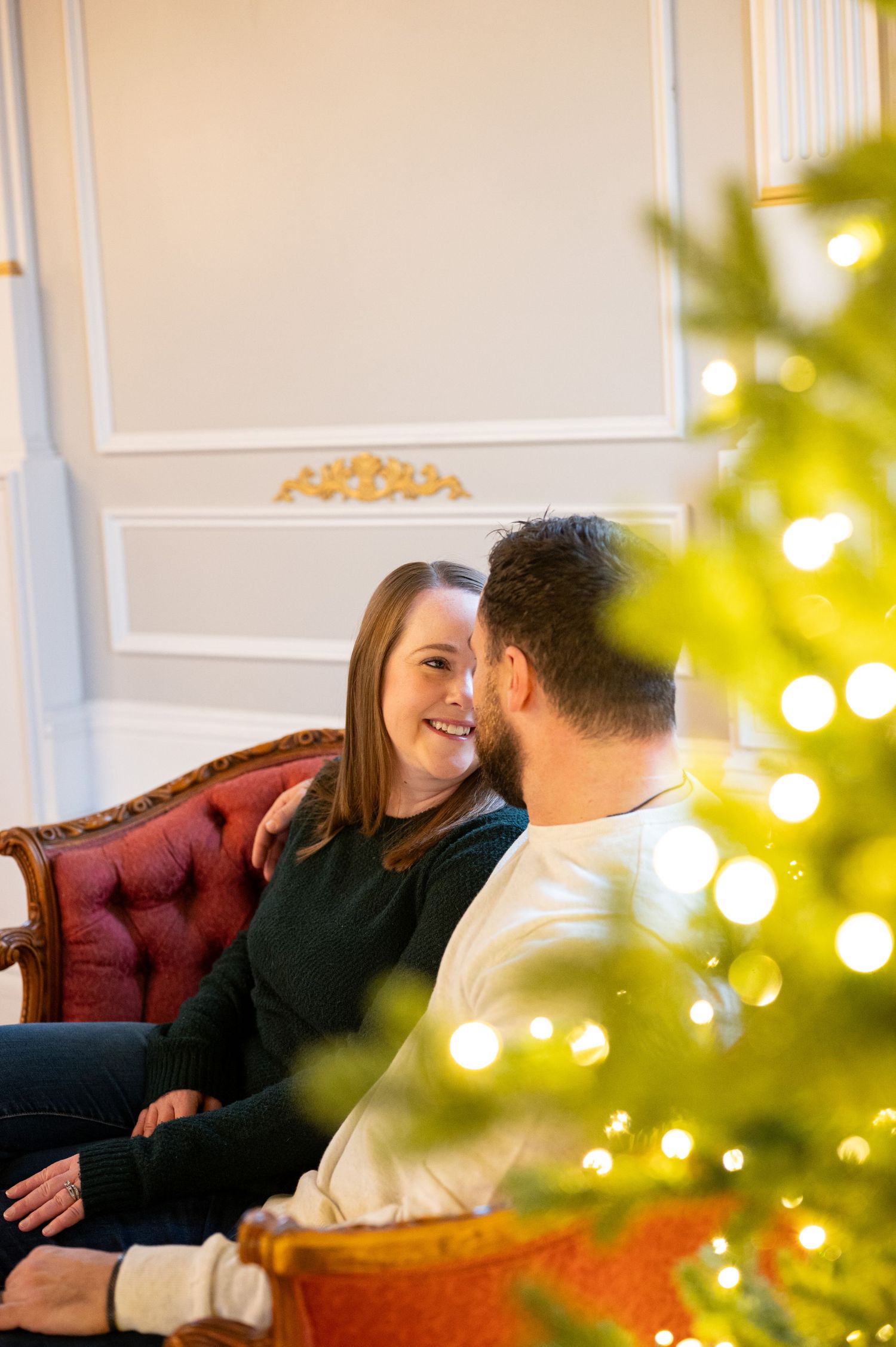 A couple shares a tender moment on a red velvet couch with a lit Christmas tree nearby. Family session. Portrait photography. Oregon.