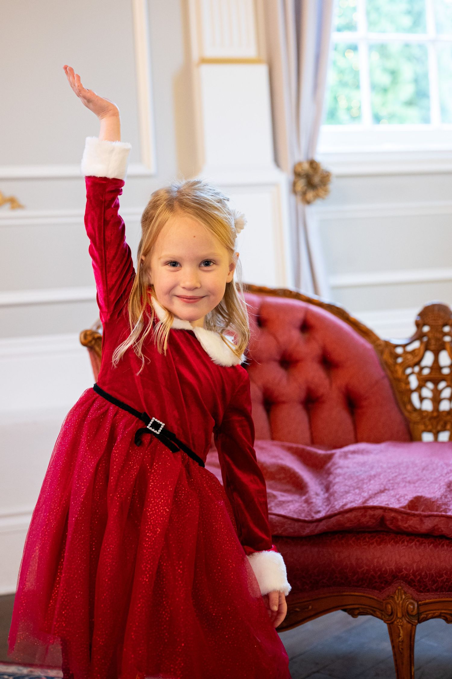 A small child in a flowing red holiday dress twirls and dances near a vintage red sofa. Family session. Portrait photography. Oregon.