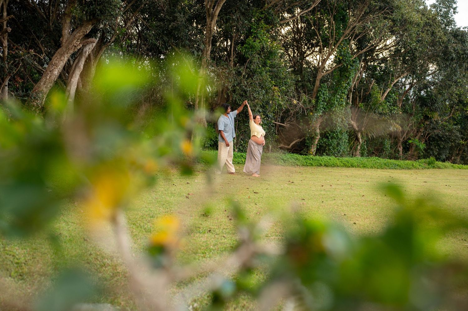 Couple walking hand in hand through a lush green park surrounded by tall trees during golden hour.