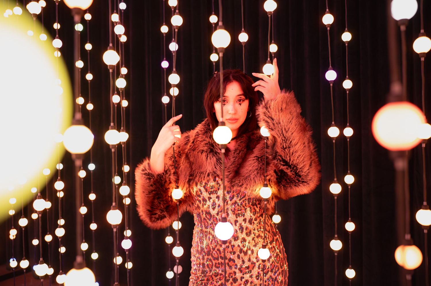 Figure poses under hanging light installation with warm ambient glow. Hopscotch. Portrait Slam.