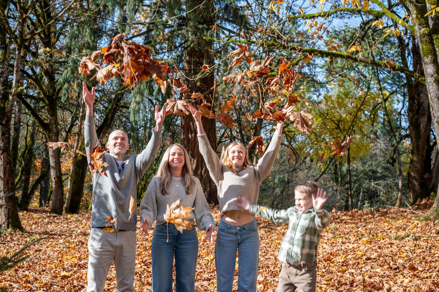 Family members jump with joy among fallen autumn leaves in a wooded park setting. Family session. Portrait photography. Oregon.
