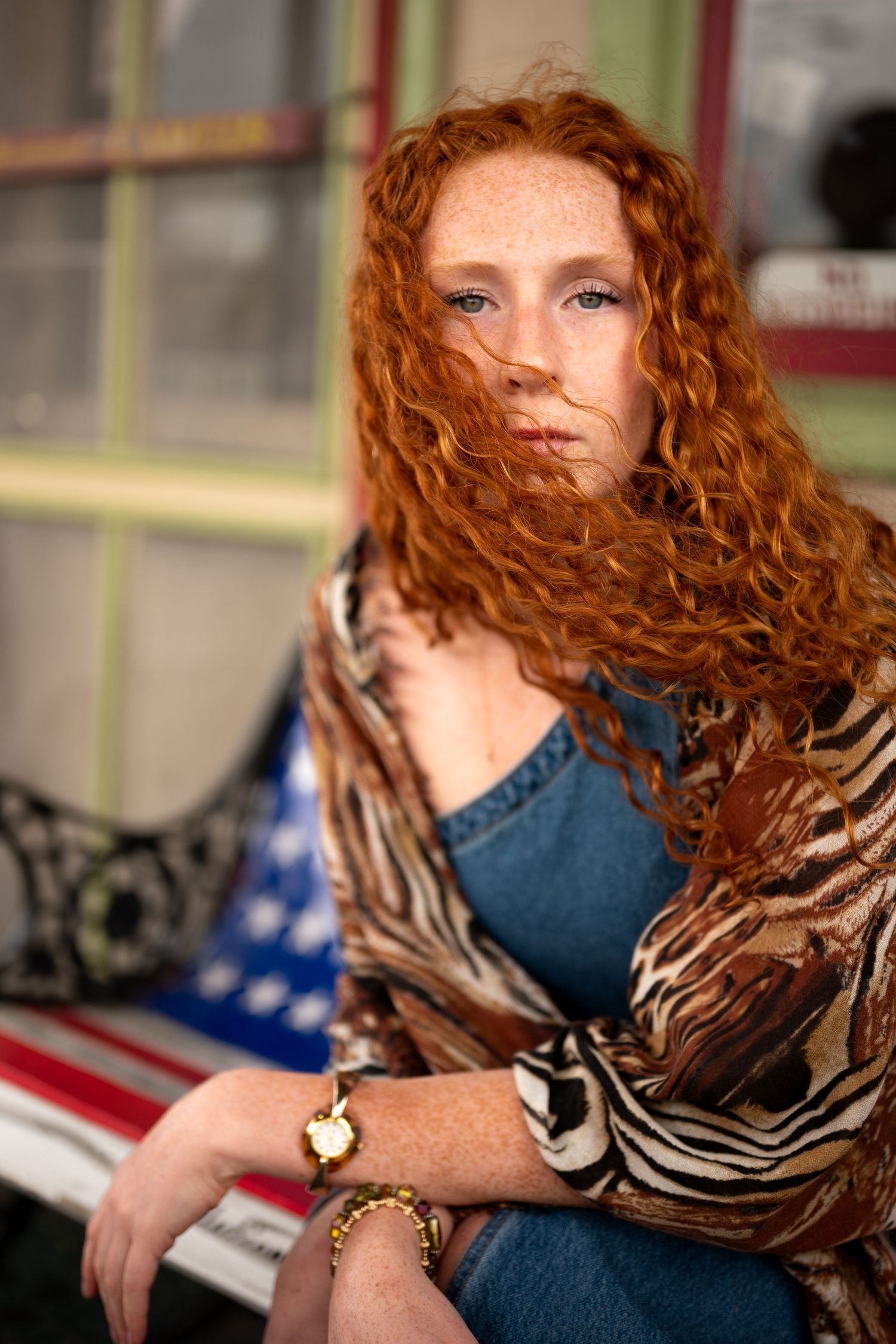 Portrait of a woman with vibrant curly red hair wearing a patterned wrap and denim against an outdoor background.