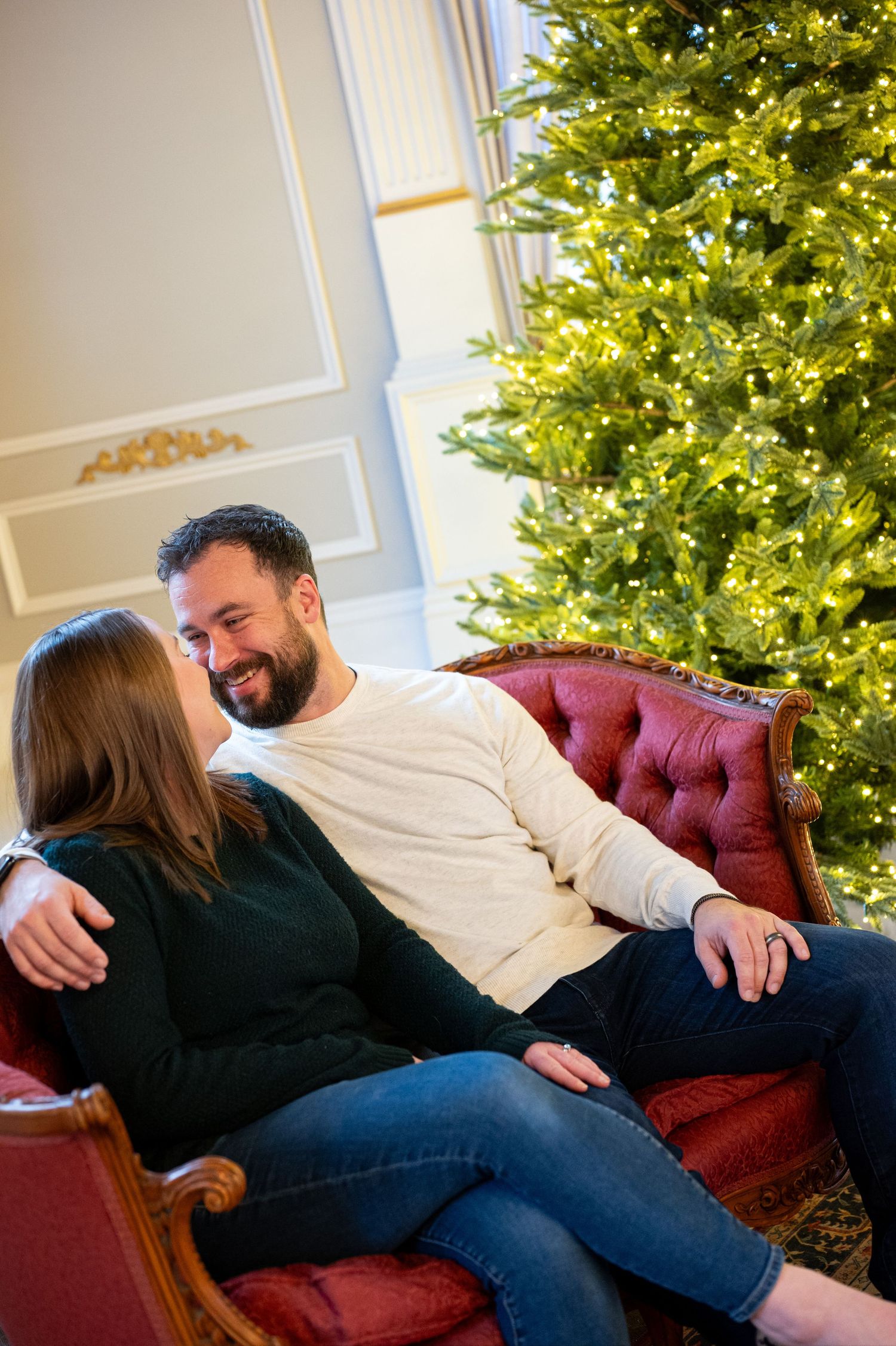 A couple lounges casually on a red vintage sofa with a decorated Christmas tree in the background. Family session. Portrait photography. Or