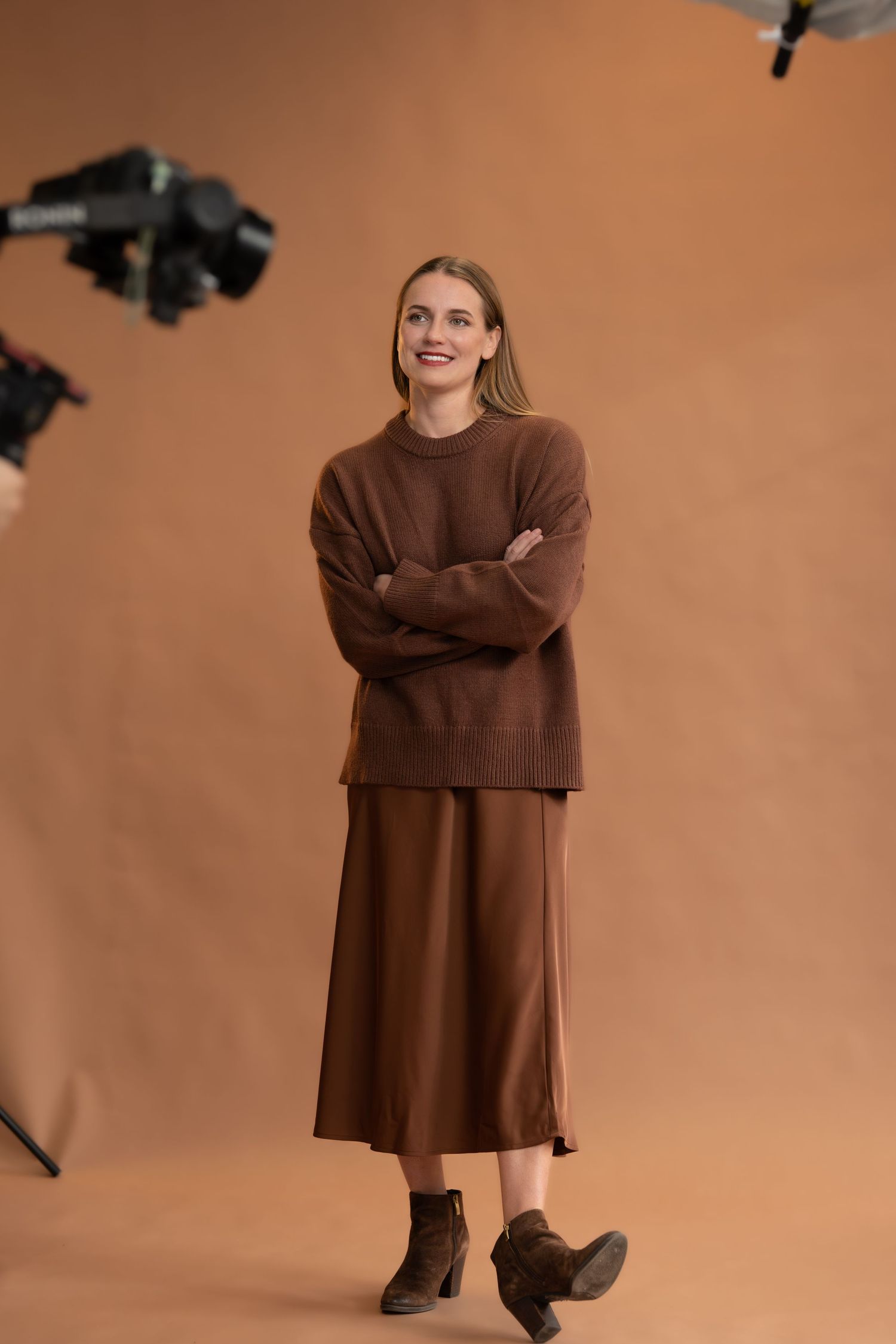 Model wearing a brown sweater and skirt ensemble poses against warm-toned backdrop during fashion photoshoot. Self portrait. Option model.