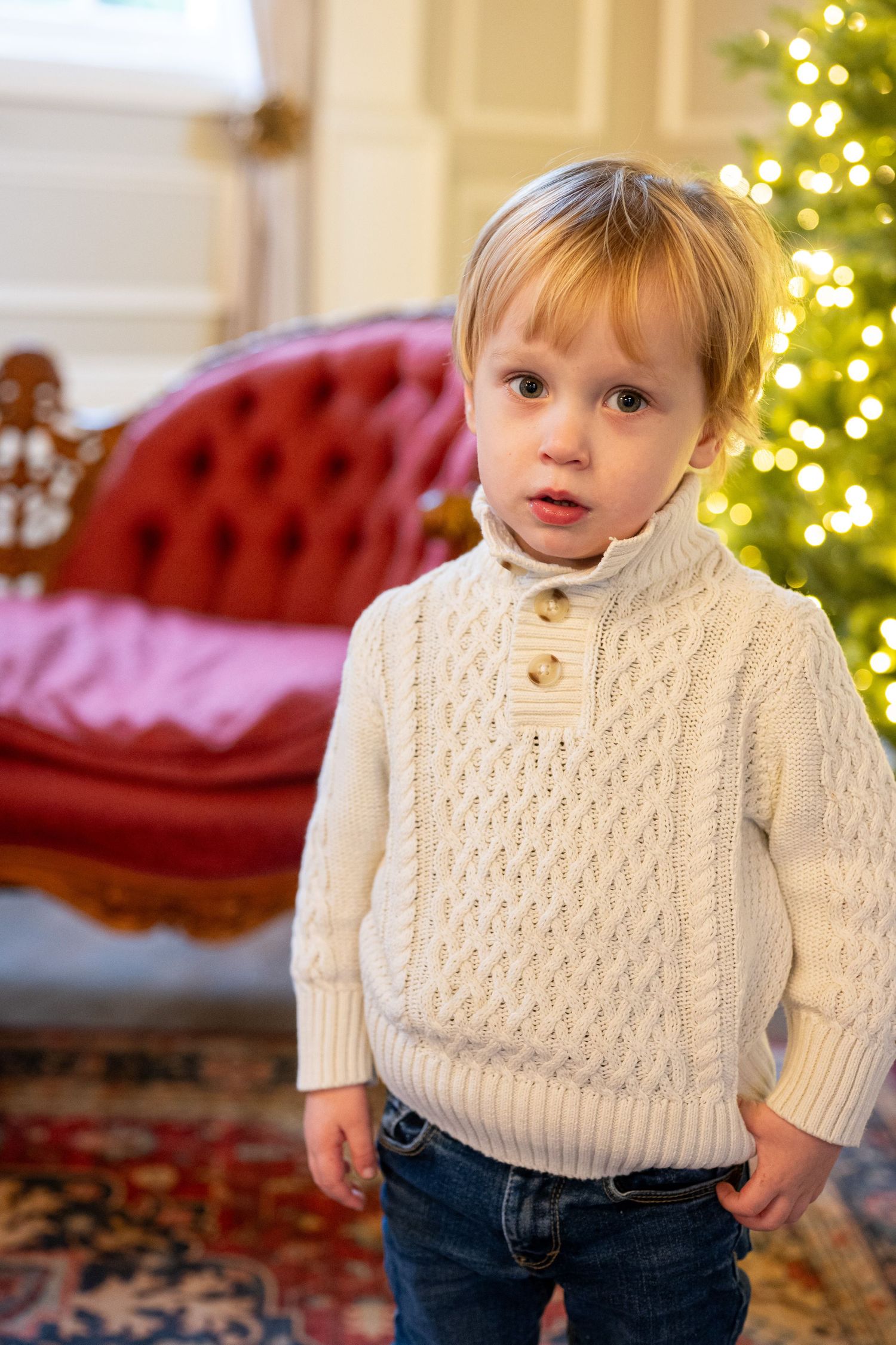 Child wearing white cable knit sweater poses in front of Christmas tree. Family session. Portrait photography. Oregon.