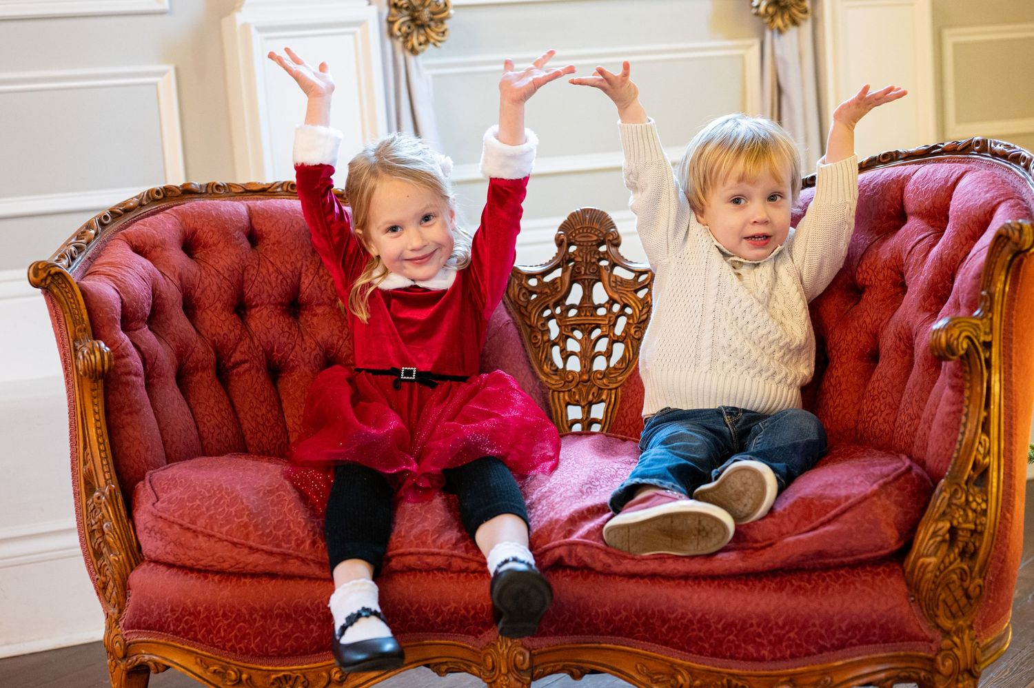 A child in white clothing sits playfully on a red velvet chaise lounge in an elegant room. Family session. Portrait photography. Oregon.