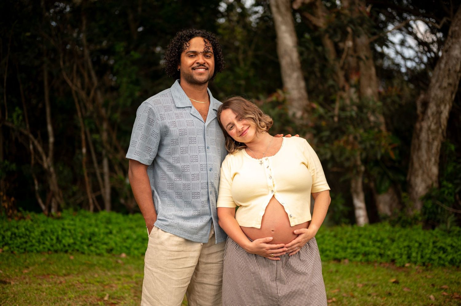 Smiling couple posing outdoors, woman in yellow top cradling baby bump, man in blue shirt, trees in background.