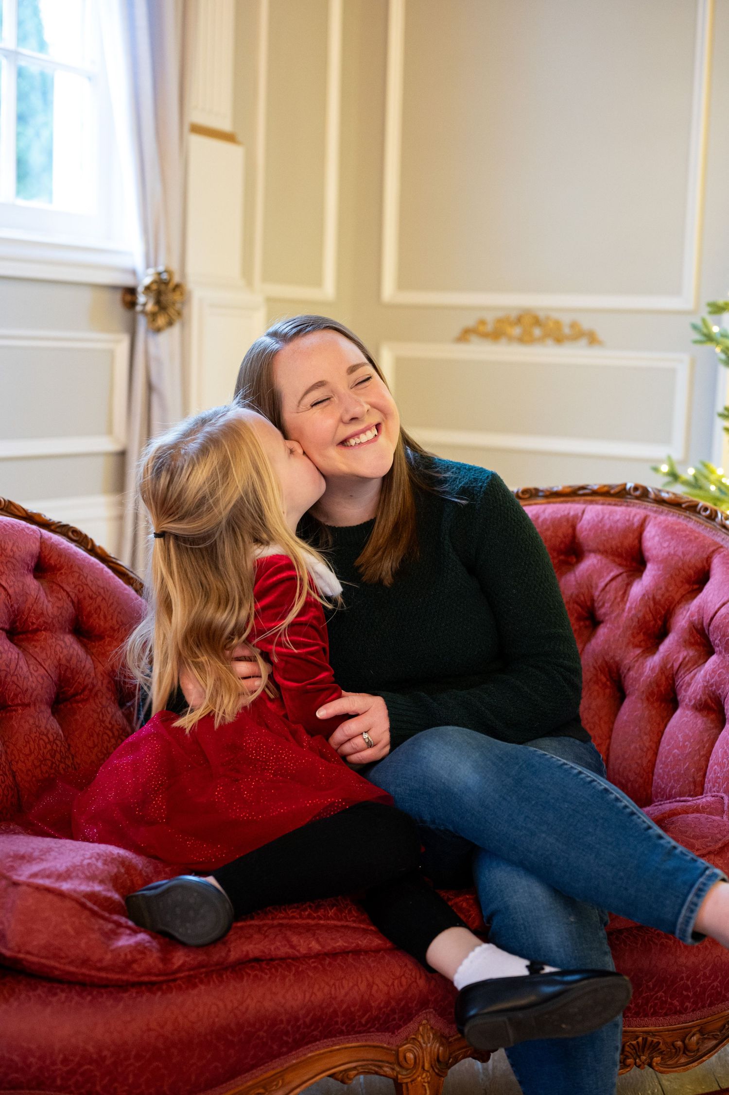Two people share a laugh while relaxing on a red velvet sofa during a holiday photoshoot. Family session. Portrait photography. Oregon.