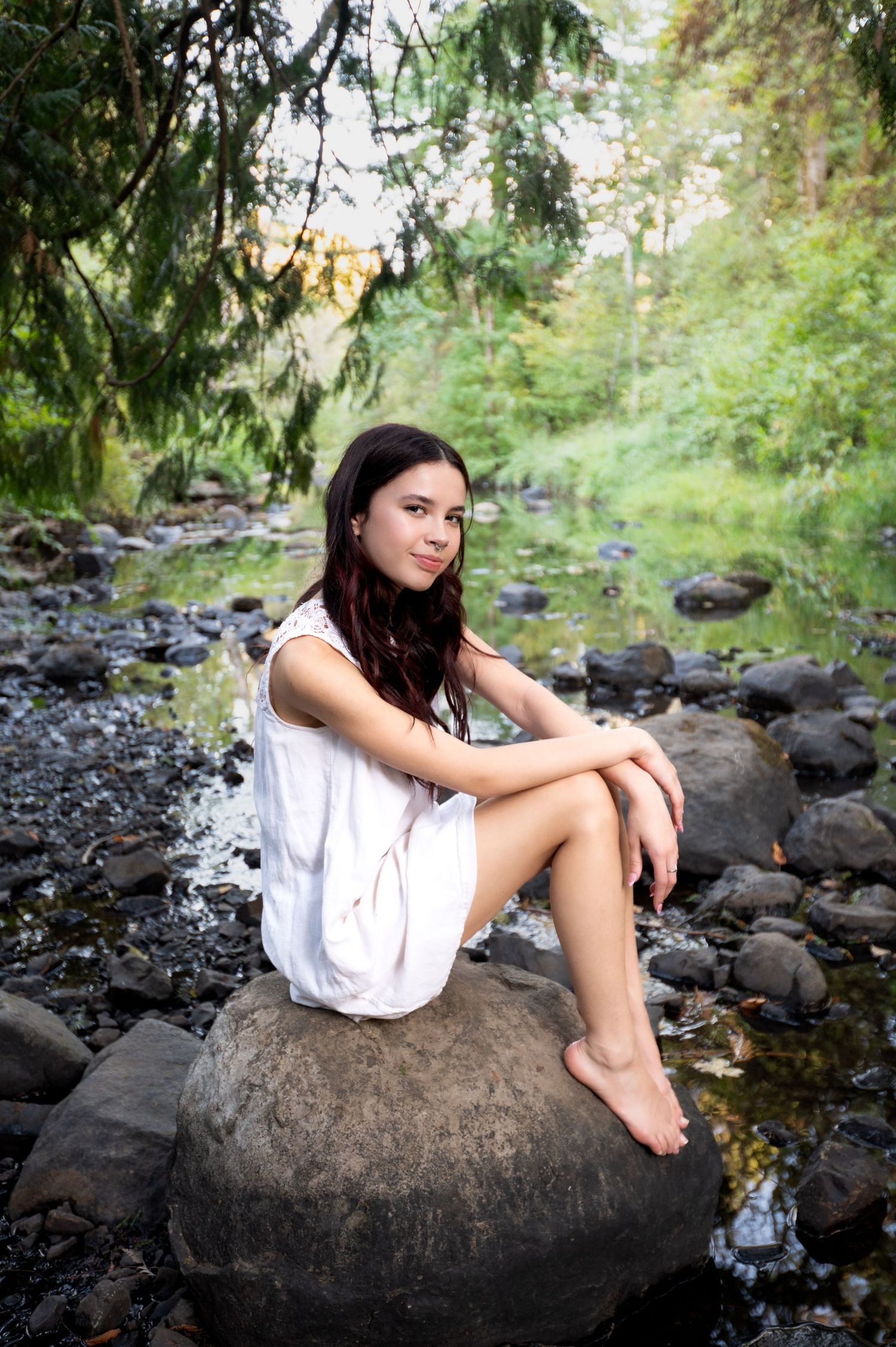 Person in white dress sitting on large rocks by a stream in a forest setting.