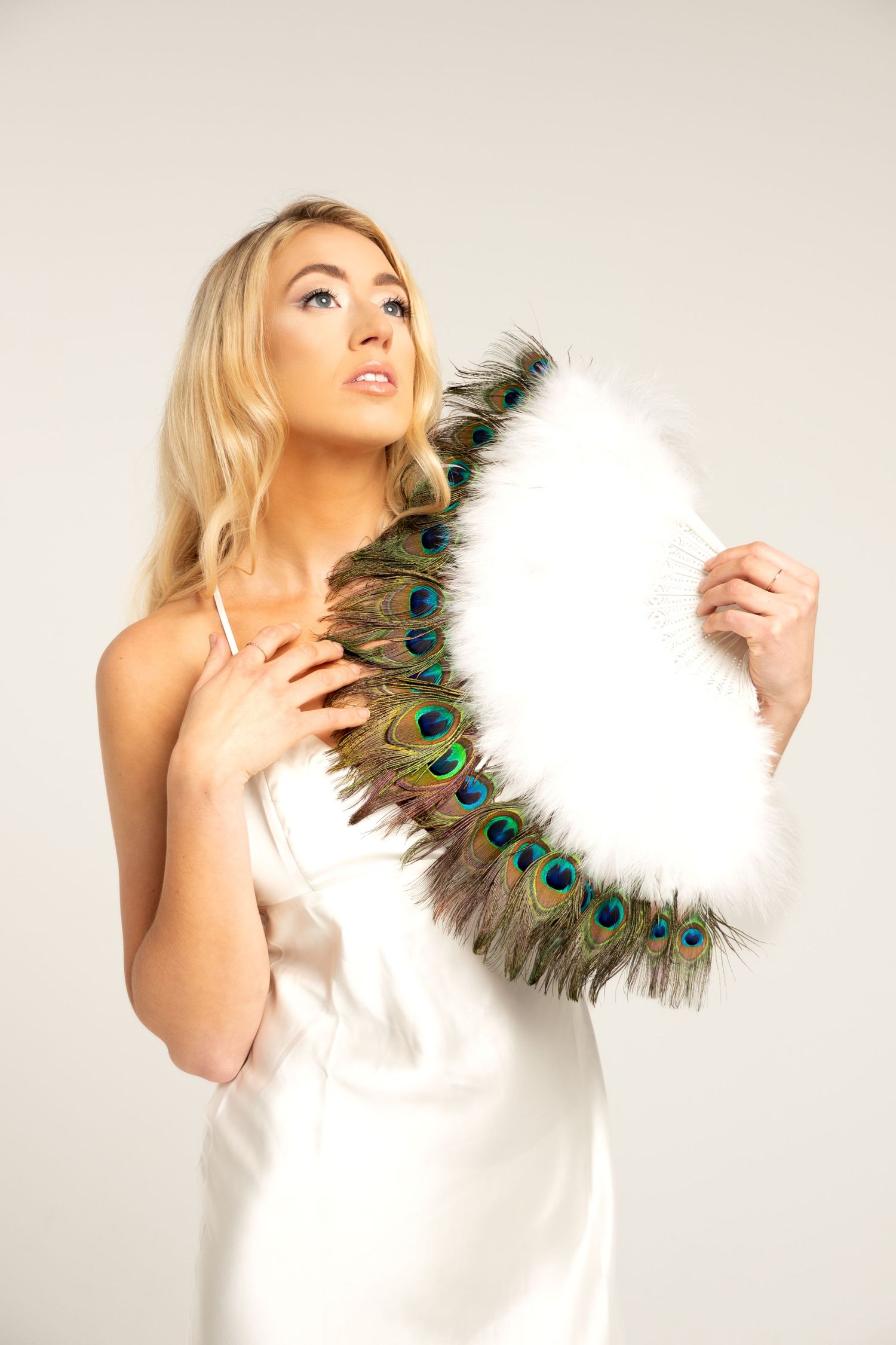 Model holds luxurious peacock feather fan while wearing flowing white dress.