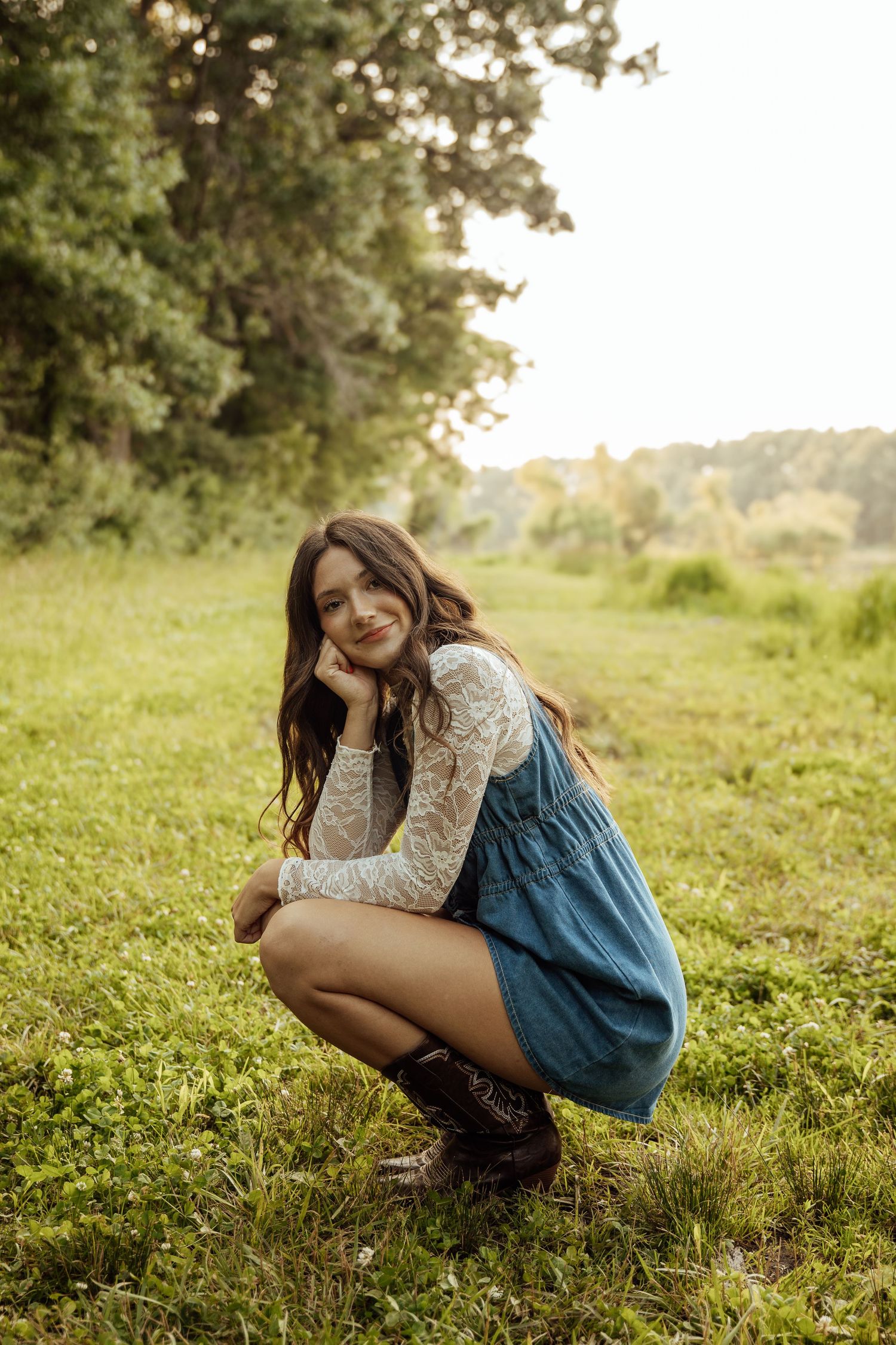 Brunette woman in denim skirt and lace top crouching and smiling in sunlit green meadow.