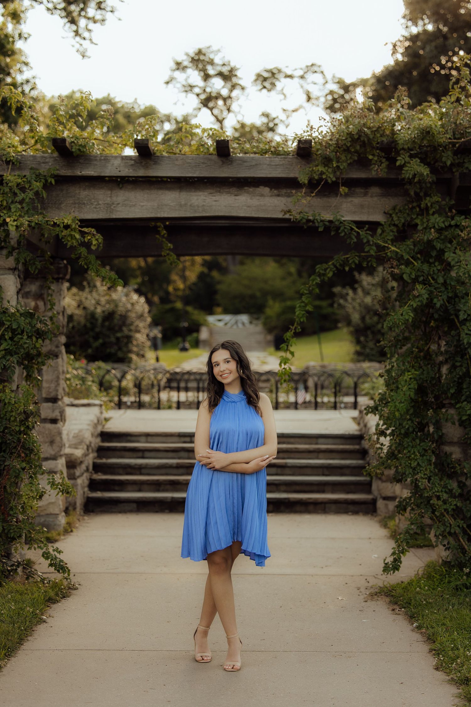 Young woman in blue halter dress posing in a garden pergola with stone steps and lush greenery.