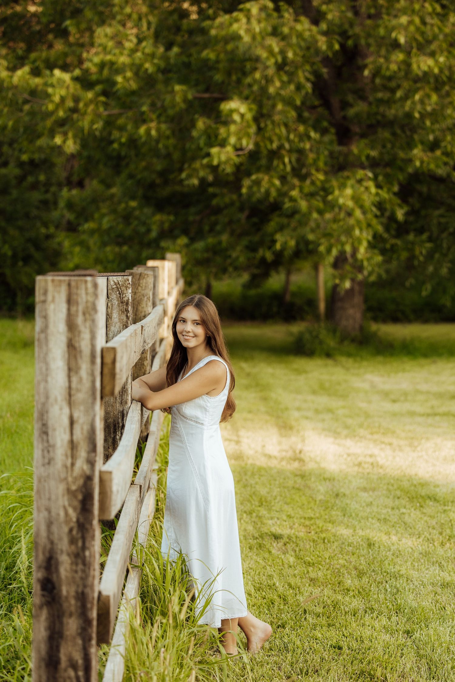 Young woman in white dress leaning on rustic wooden fence surrounded by lush green trees and grass.