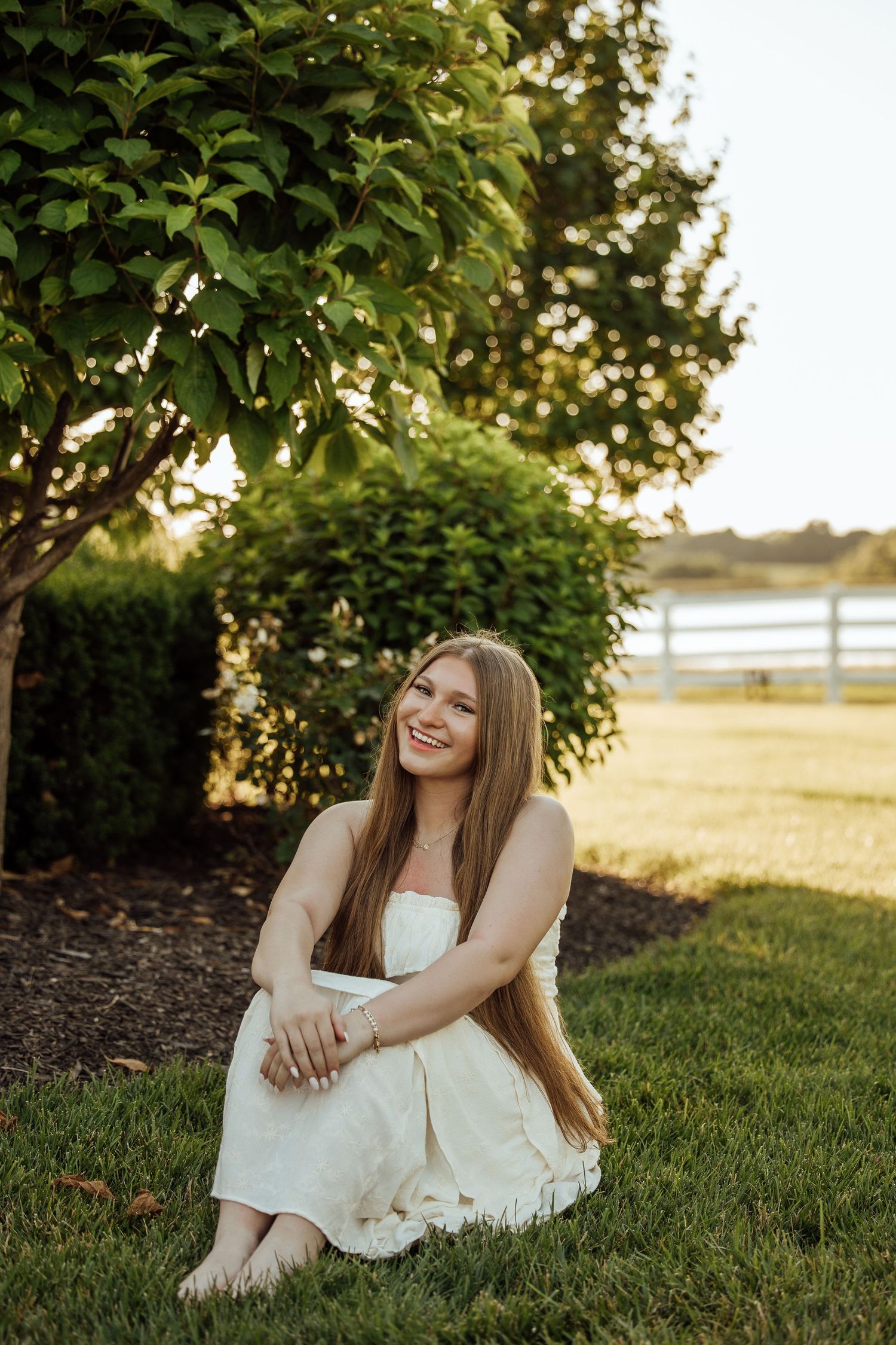 Young woman in white dress sitting on green grass, smiling, with trees and golden hour light in background.