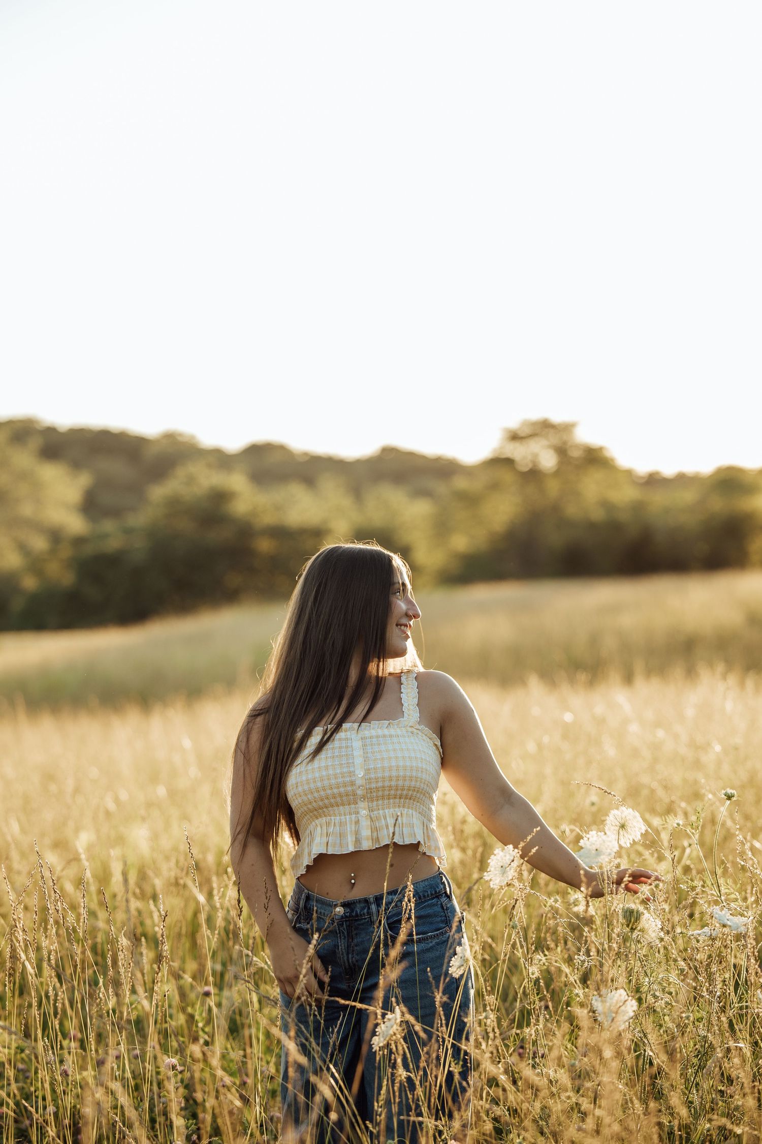 Young woman in white crop top and jeans smiling in golden field at sunset, holding wildflowers.