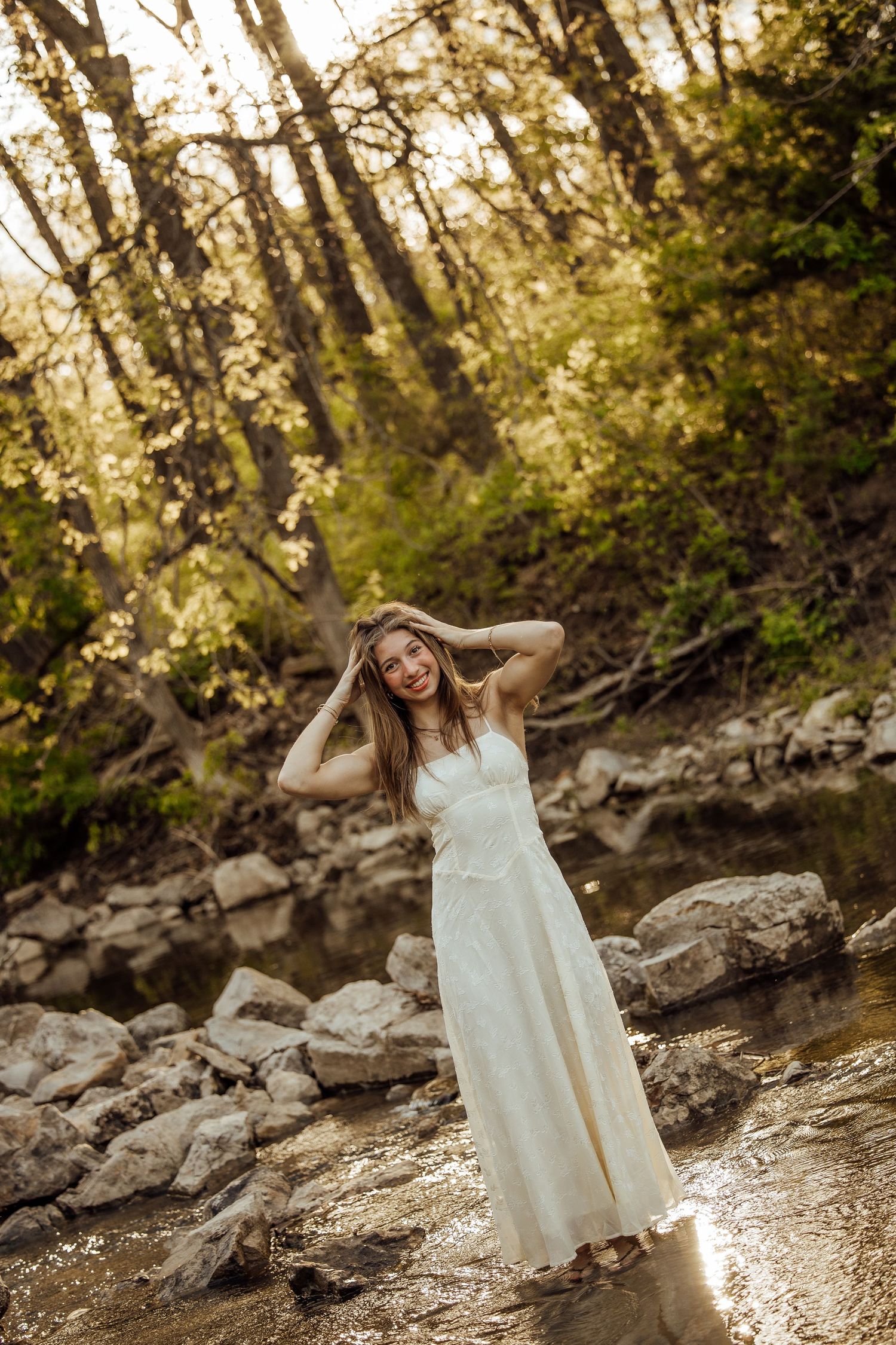 Joyful bride in white gown standing in rocky creek surrounded by golden sunlit forest trees.