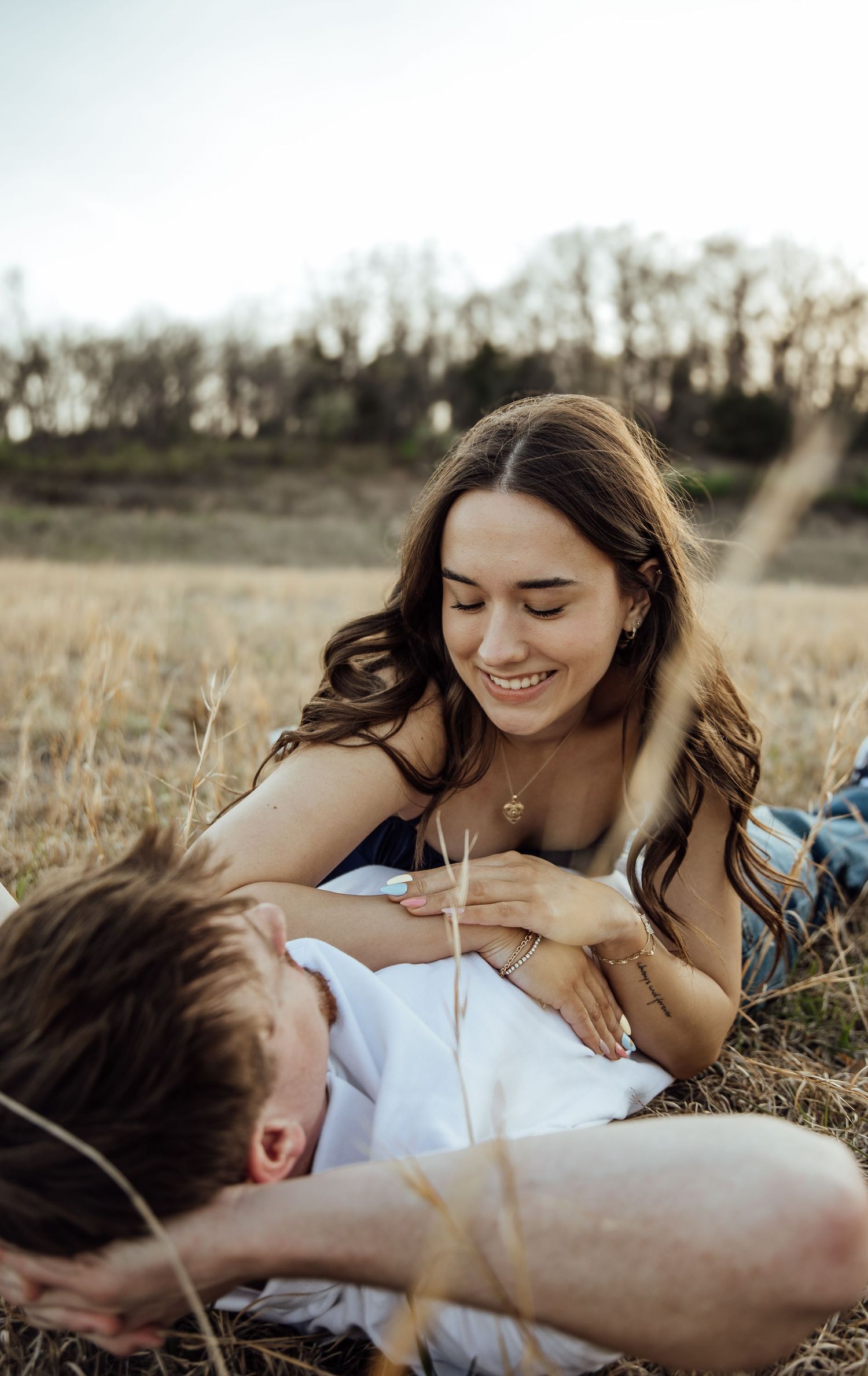 A smiling woman with long brown hair leans over a man lying in a golden field during a romantic outdoor couples session.