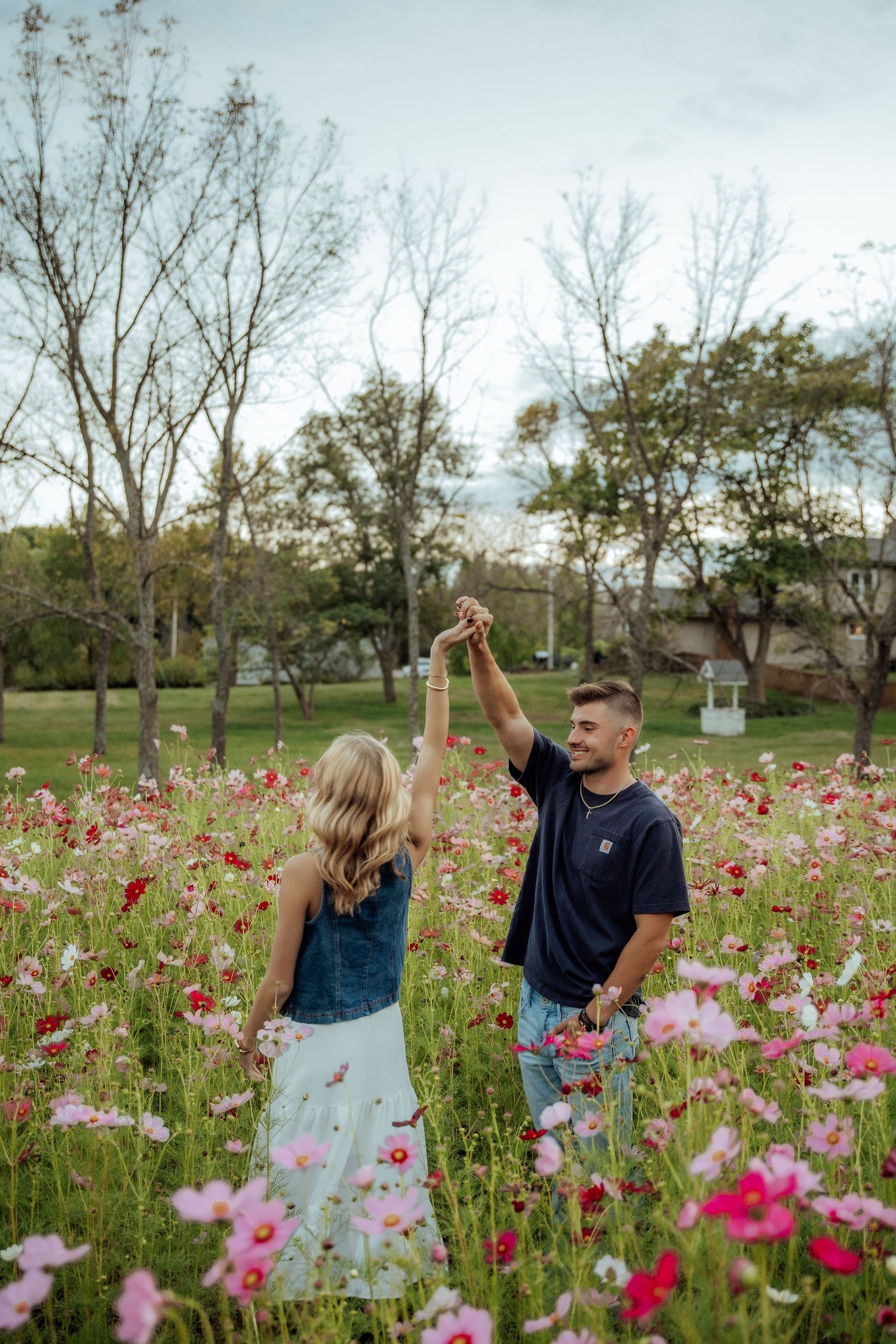 Couple dancing in vibrant pink flower field, woman in denim top and skirt, man in navy shirt holding flowers.