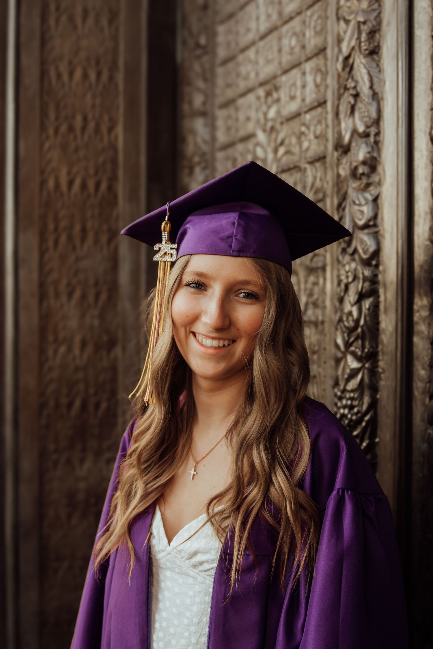 Smiling graduate in purple cap and gown with 2025 tassel standing by ornate carved doors.