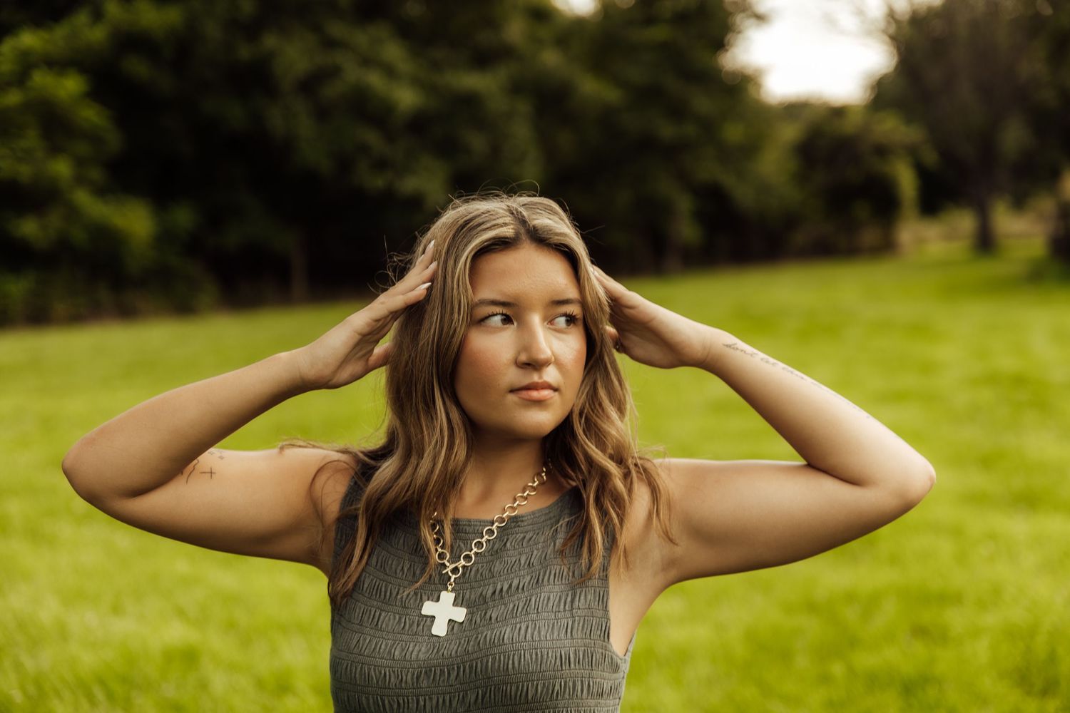 Young woman in gray top with cross necklace poses outdoors in green park setting, hands in hair.