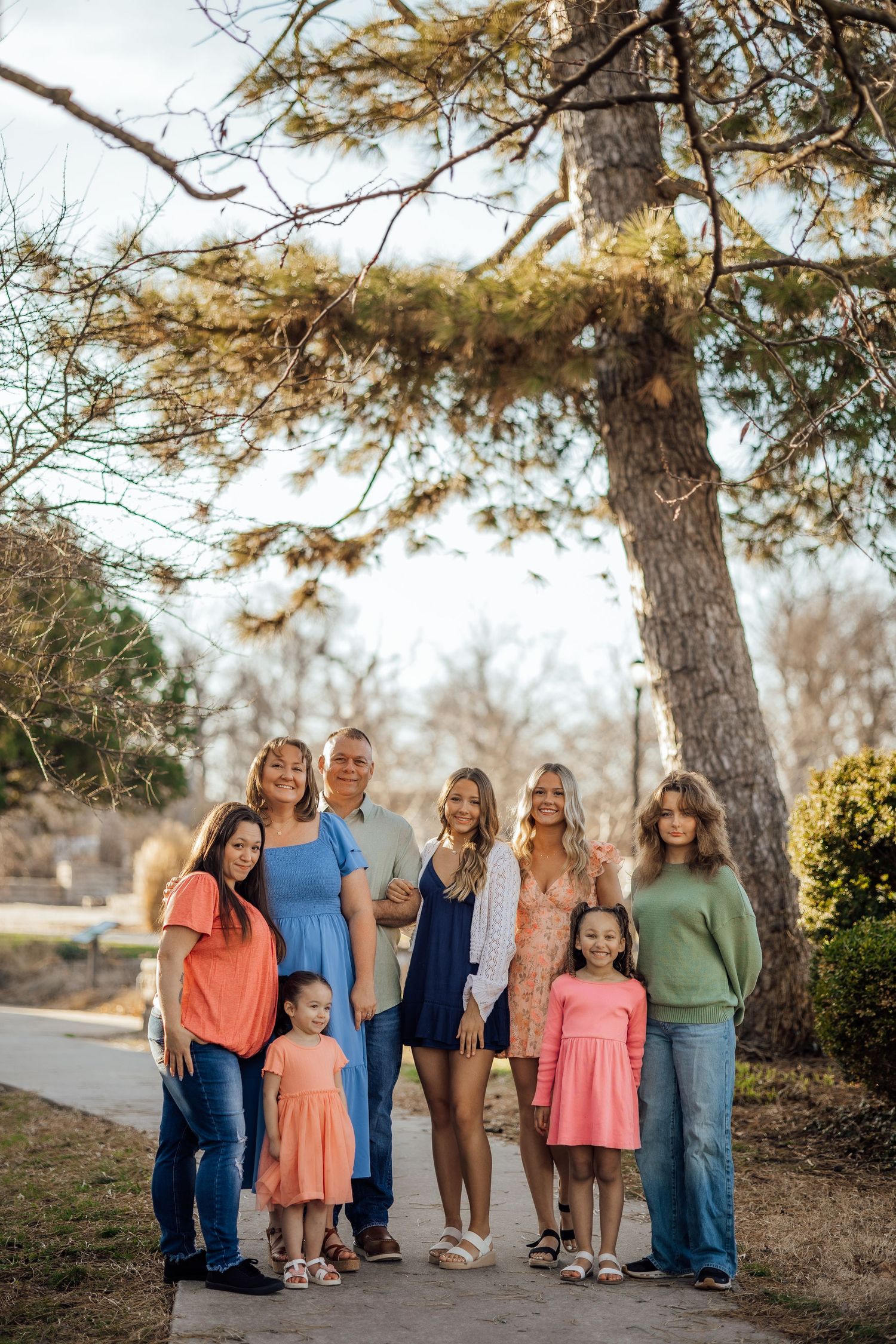 Extended family of eight posing together under a large pine tree during an outdoor family portrait session.