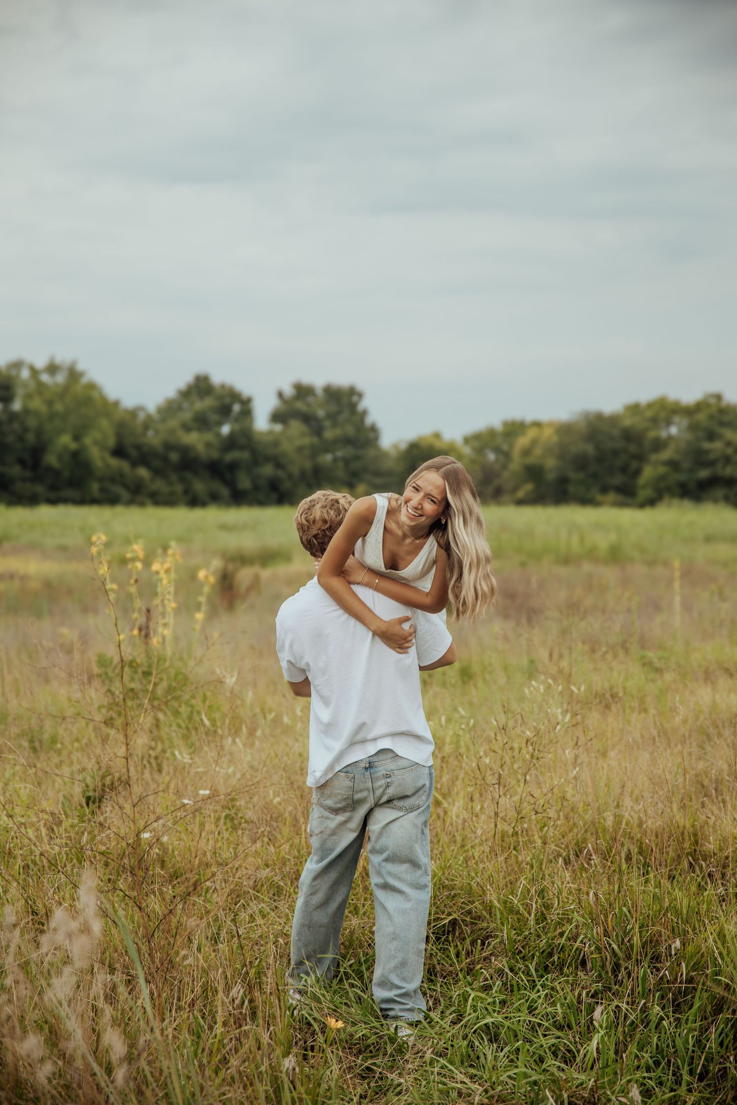 A couple embraces and laughs together in a scenic grassy field with trees in the background.