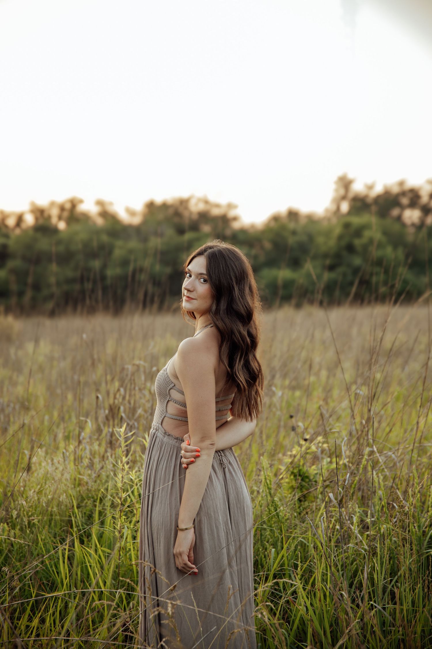 Brunette woman in gray maxi dress standing in tall grass field during golden hour sunset.