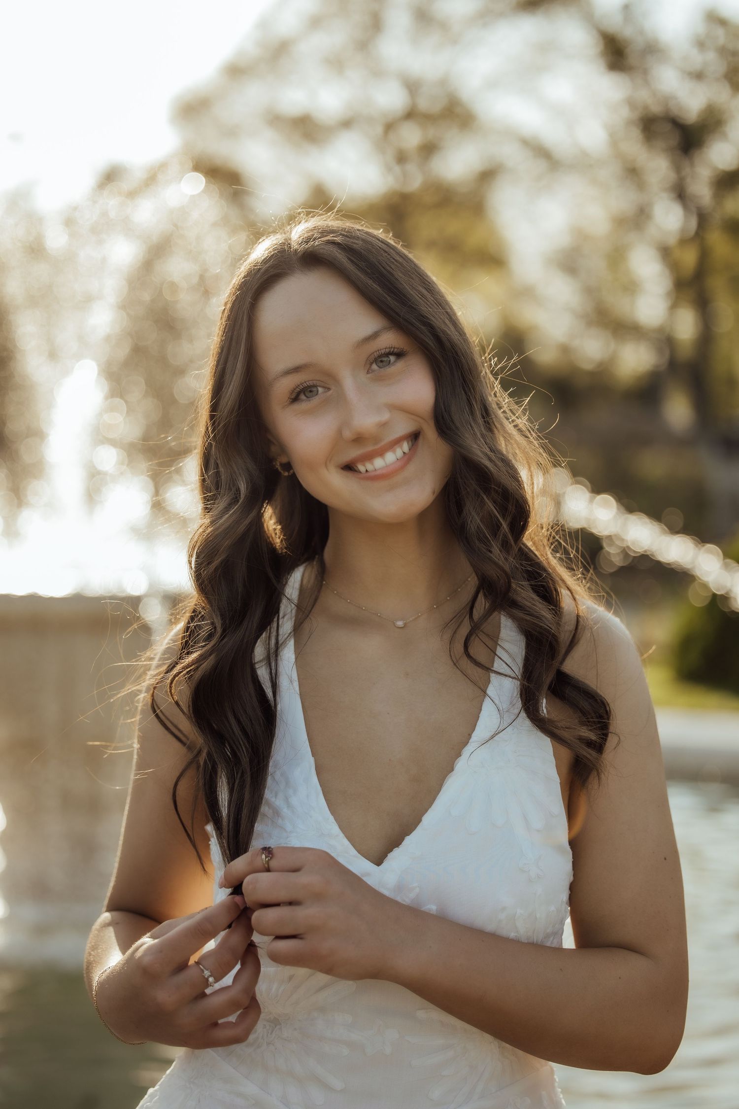 Brunette woman in white v-neck dress smiles in golden sunlight near a fountain.