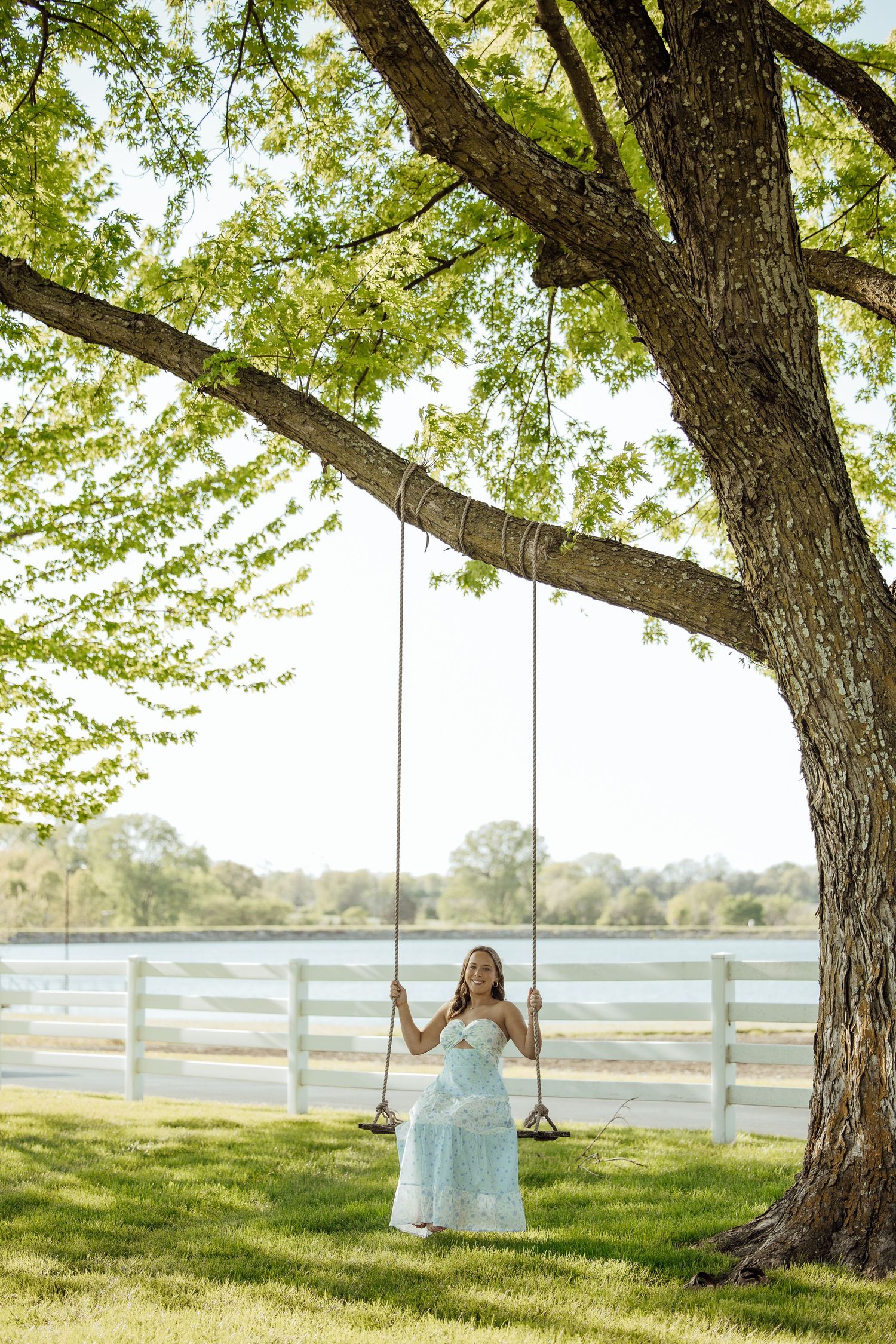 Young woman in blue floral dress sitting on rope swing under large tree by lakeside.