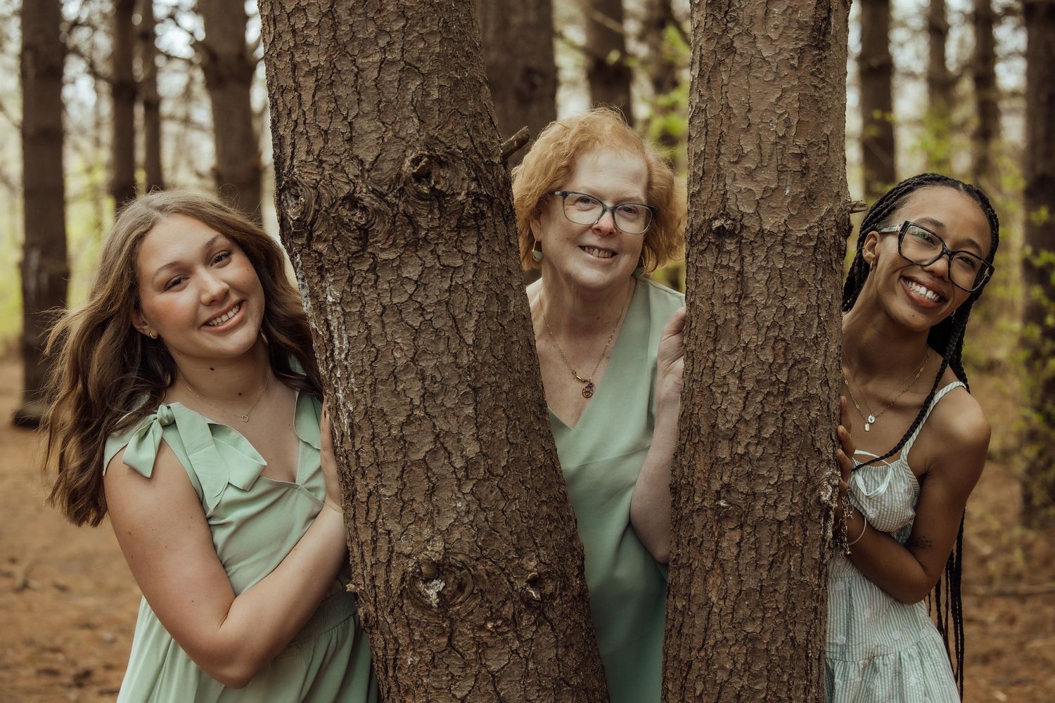 Grandmother and two granddaughters playfully peeking around pine trees during an outdoor family photo session.