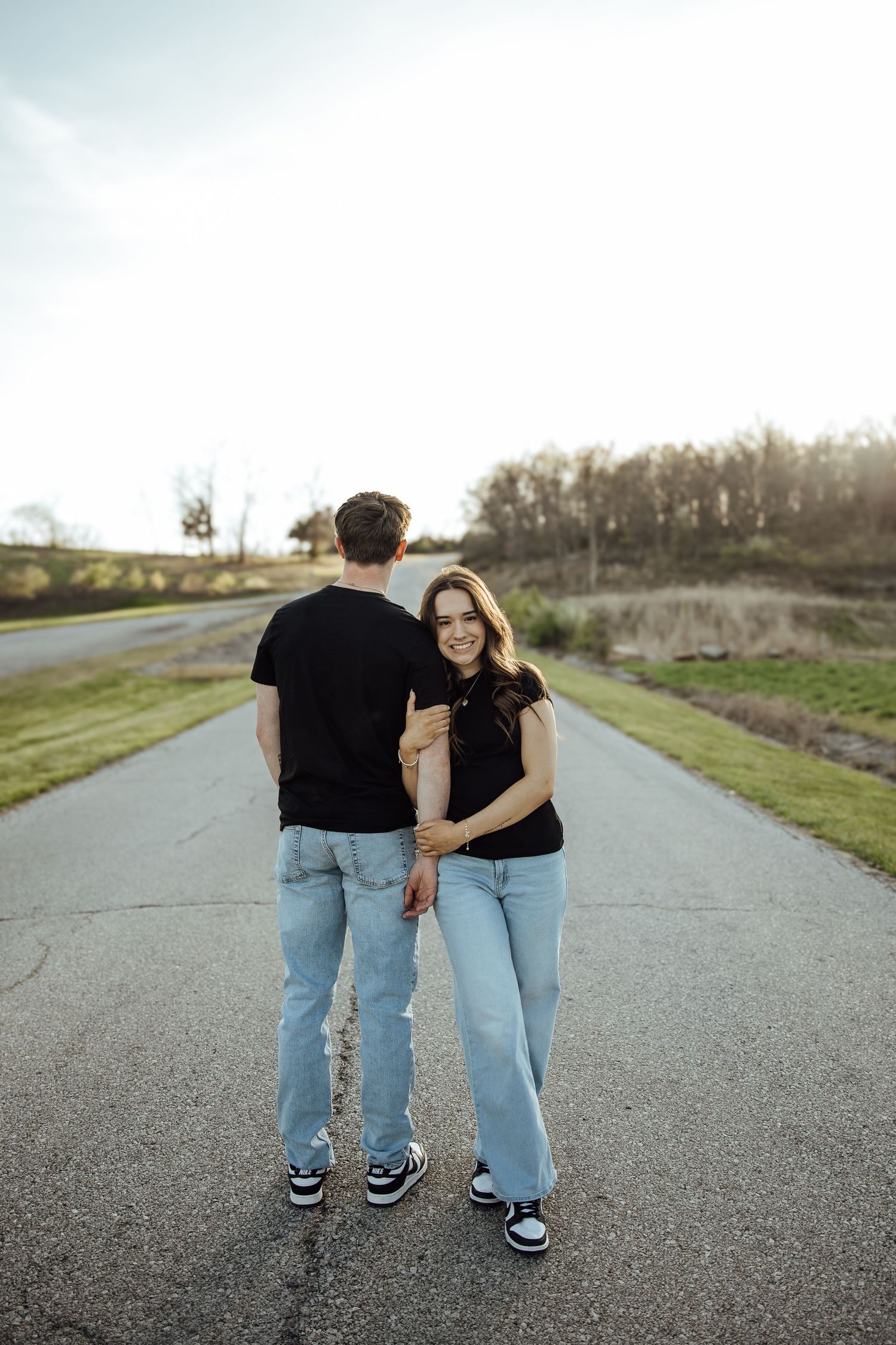 A smiling young woman in black top and jeans holds hands with her partner on a quiet rural road at sunset.
