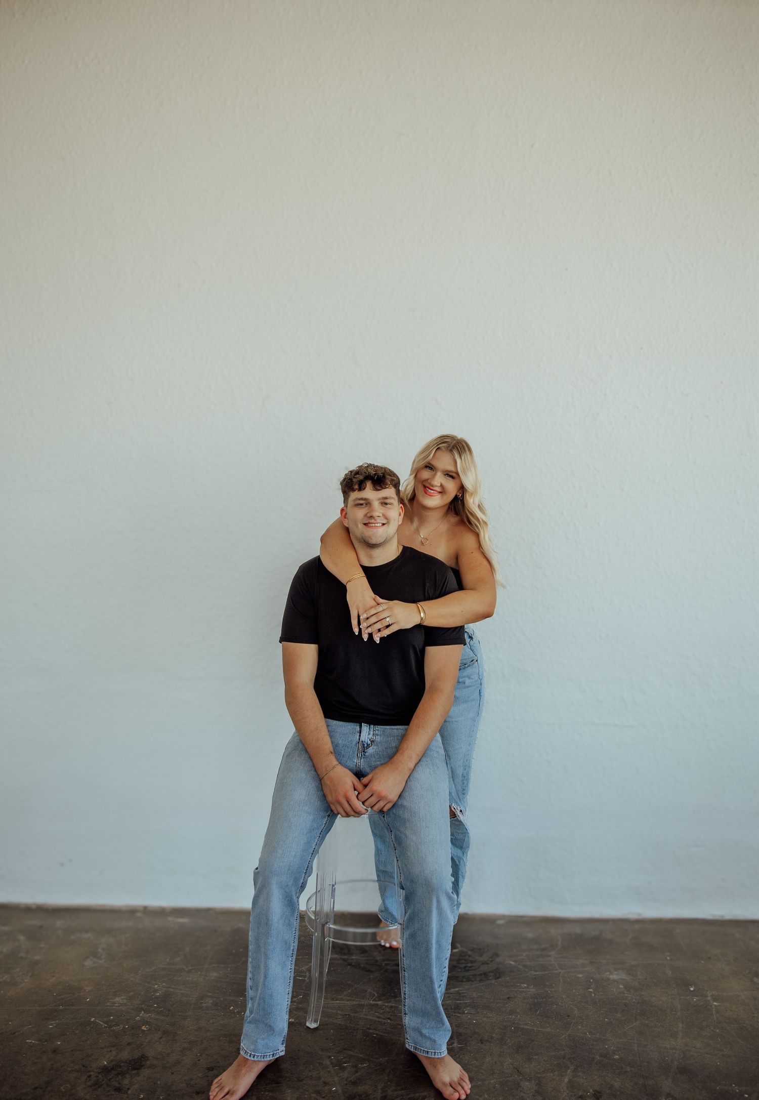 Couple posing against white wall, man seated on clear chair with woman hugging him from behind, both in jeans.