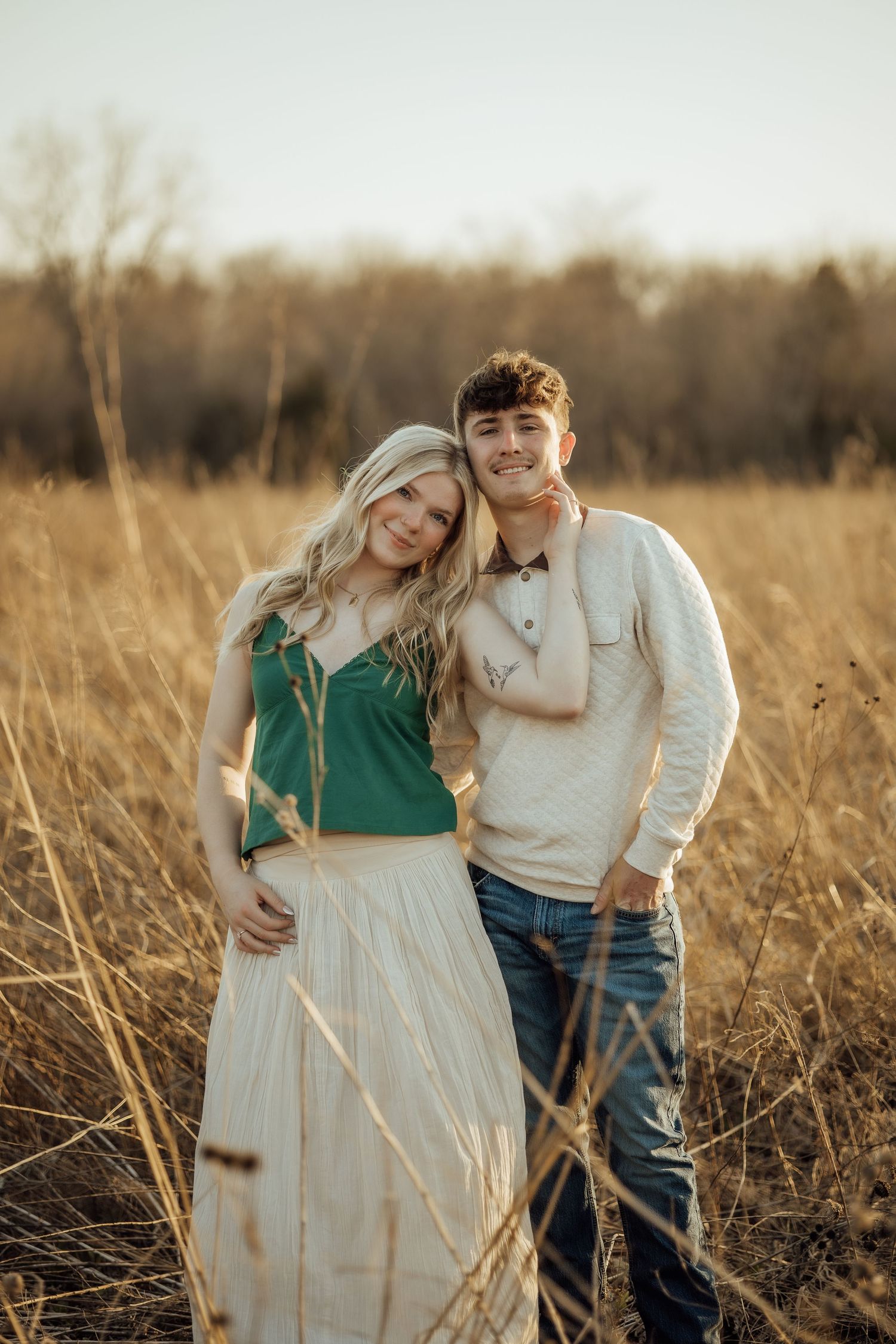Young couple embracing in golden field at sunset, woman in green top and white skirt, man in cream sweater and jeans.