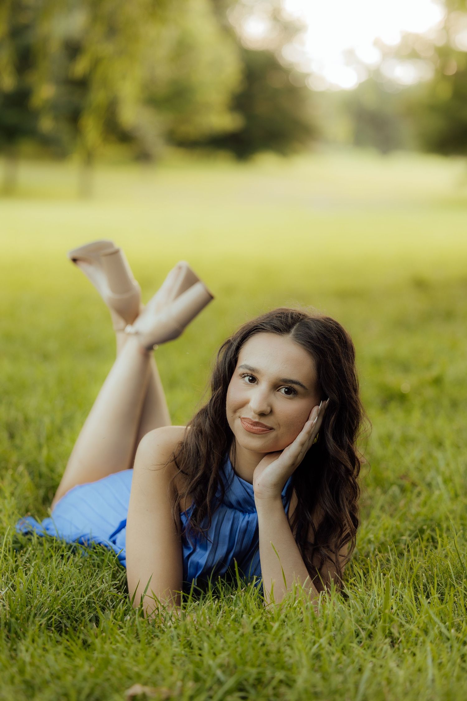 Young woman in blue dress lying on green grass in a park, resting her head on her hand, smiling.