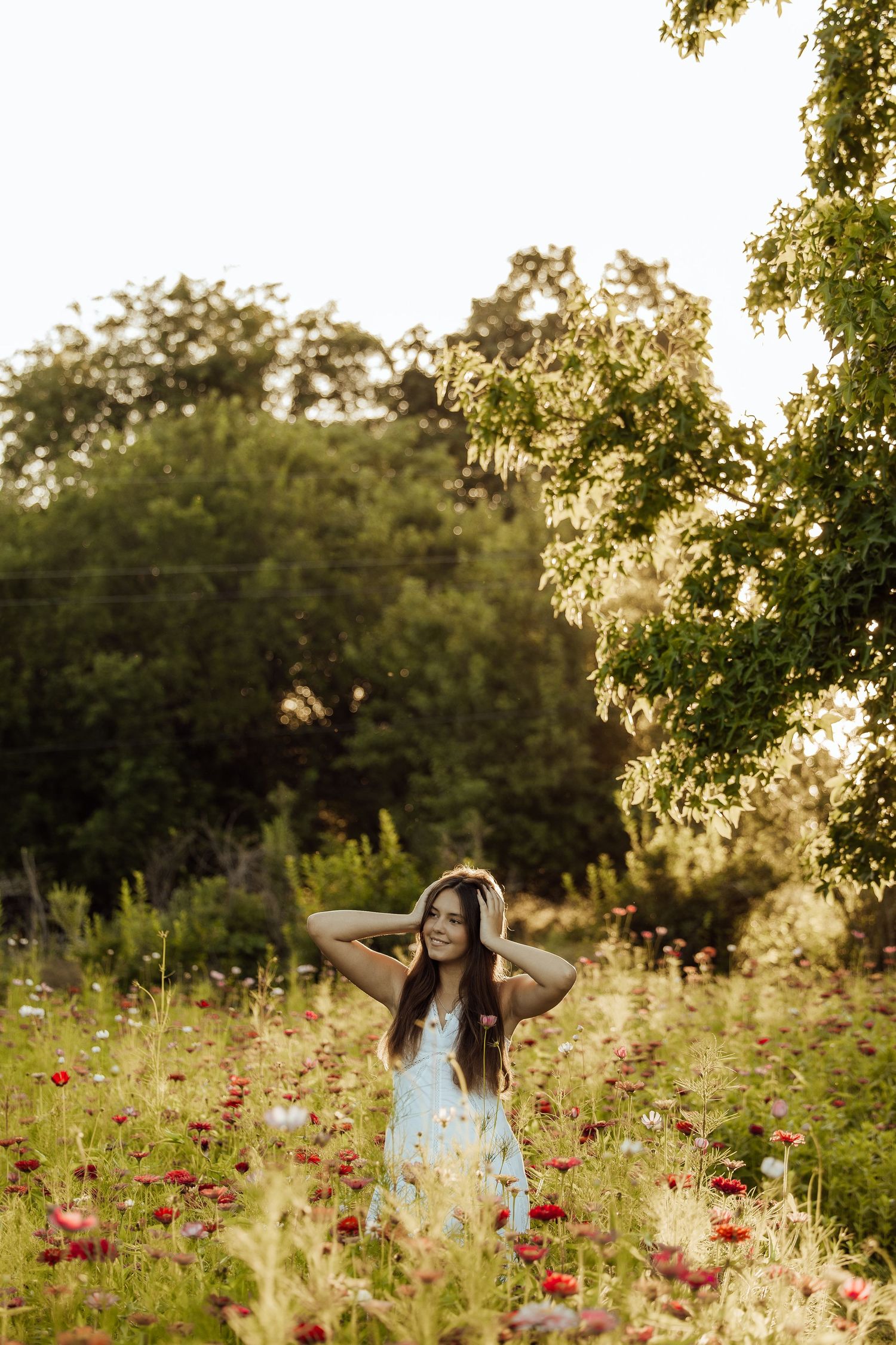 Smiling brunette woman in floral dress standing in a red poppy field surrounded by green trees in warm sunlight.