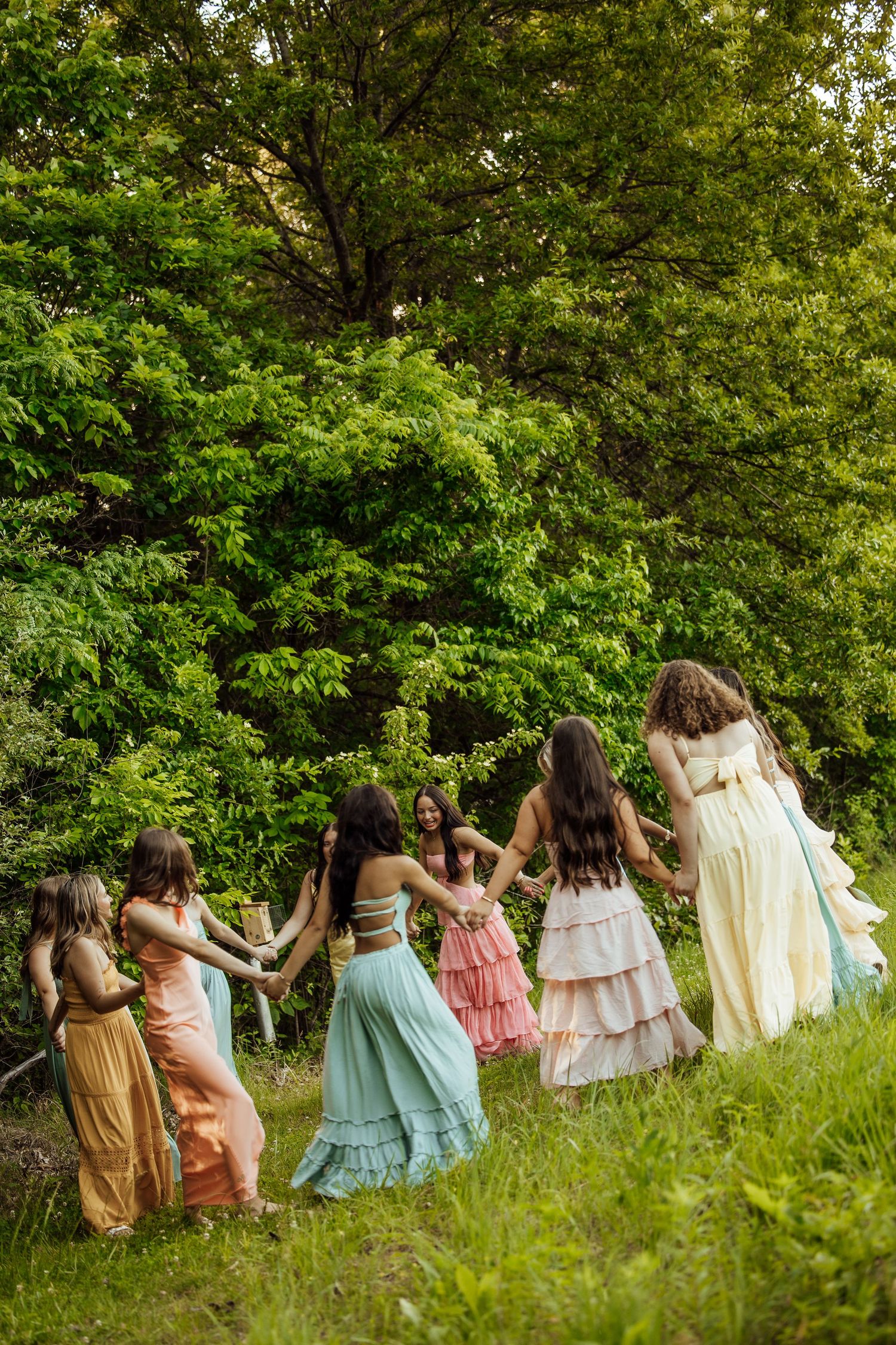 Group of women in colorful flowing dresses holding hands in circle near lush green trees.