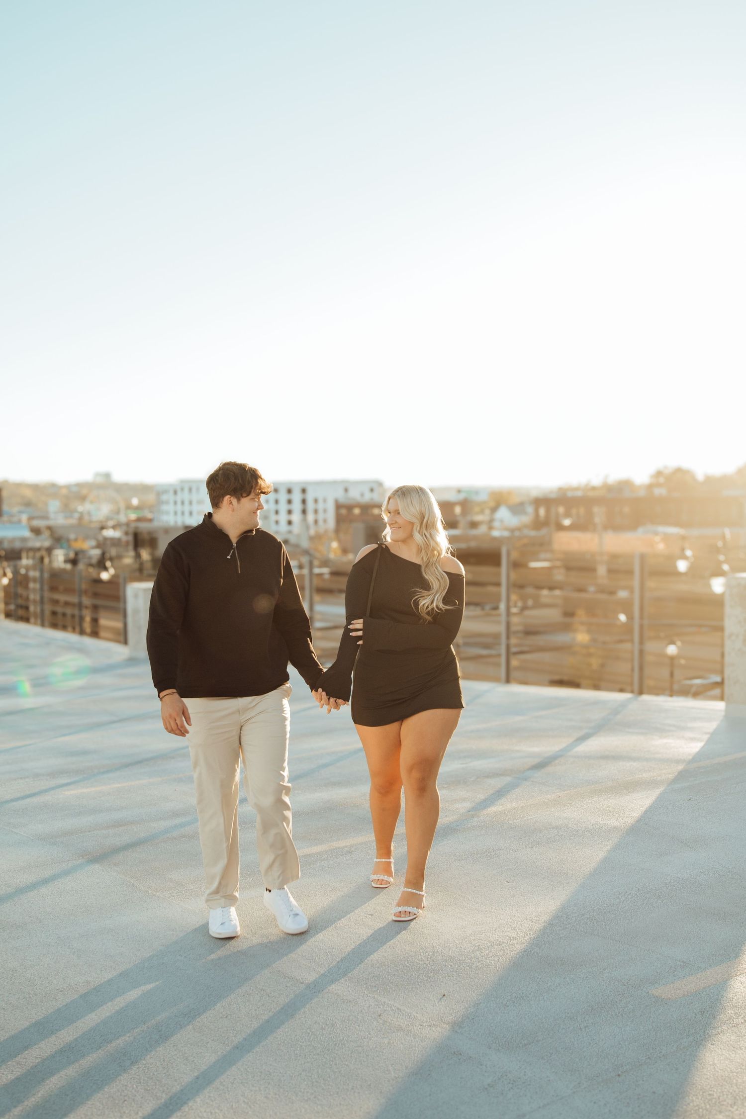 A couple holds hands and walks on a rooftop at golden hour with a sunny cityscape in the background.