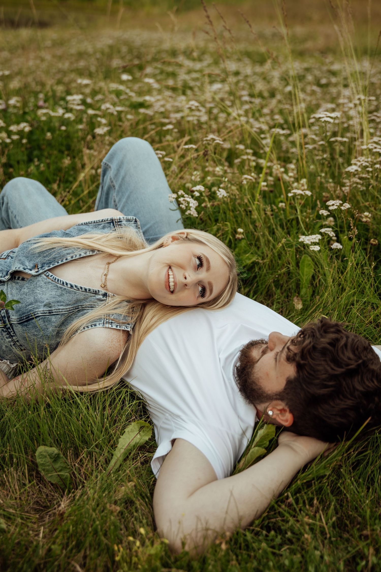 A happy couple lies in a flower-filled meadow, smiling and gazing at each other lovingly.